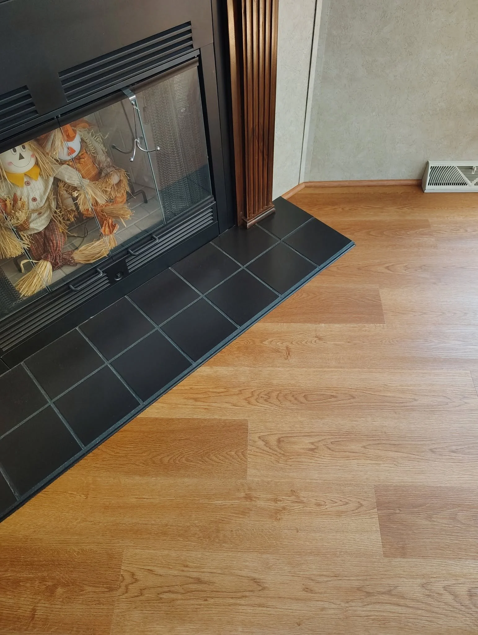 The image shows a corner of a room with a fireplace. The fireplace has a glass door with a couple of scarecrow decorations inside. To the right of the fireplace is a wooden trim, and the floor is a mix of black tiles and light-colored wood.