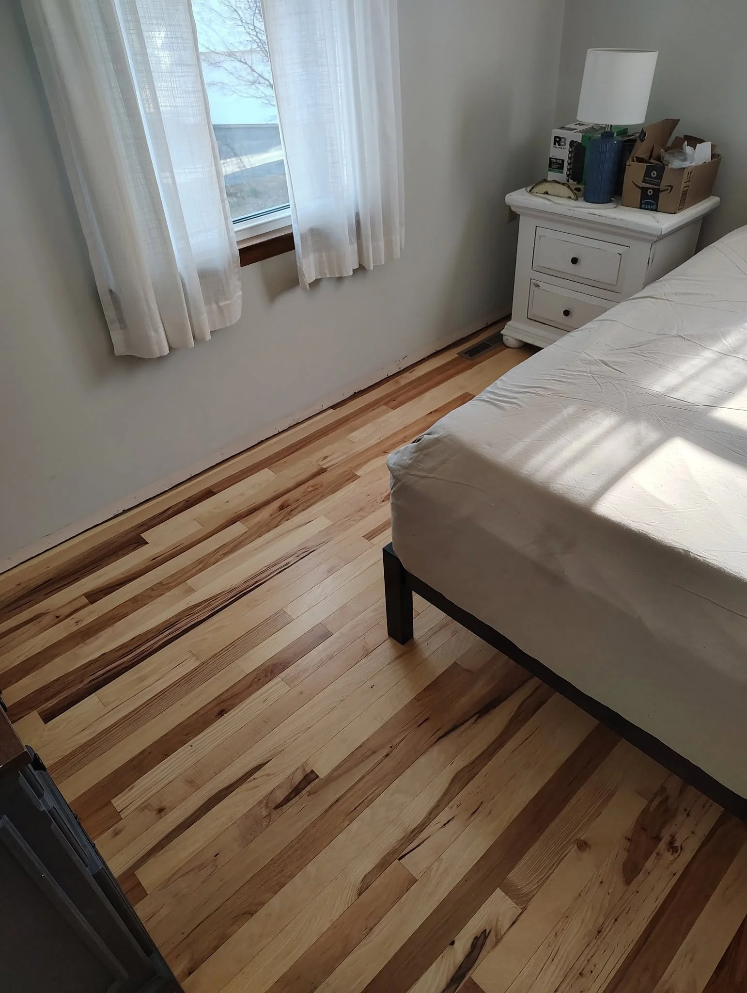 A bedroom with a wooden floor, a bed with white sheets, a white nightstand, a lamp, and some cardboard boxes on the nightstand. There is a window with white curtains and a view of the outdoors with trees.