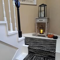 Entryway with staircase, black and white striped rug, small white table with candle inside lantern, and pumpkins on the side.
