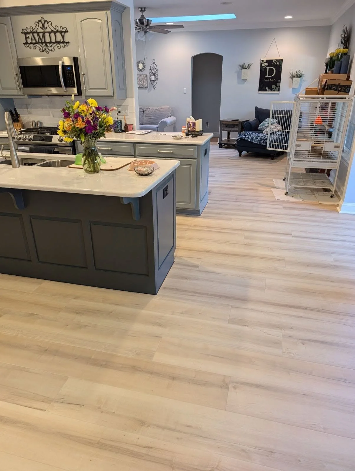 A kitchen with light-colored cabinets, a white marble countertop, a vase of colorful flowers, a microwave, and various small items. The kitchen opens into a living area with a dark sofa, wall decorations, a ceiling fan, and a hardwood floor.