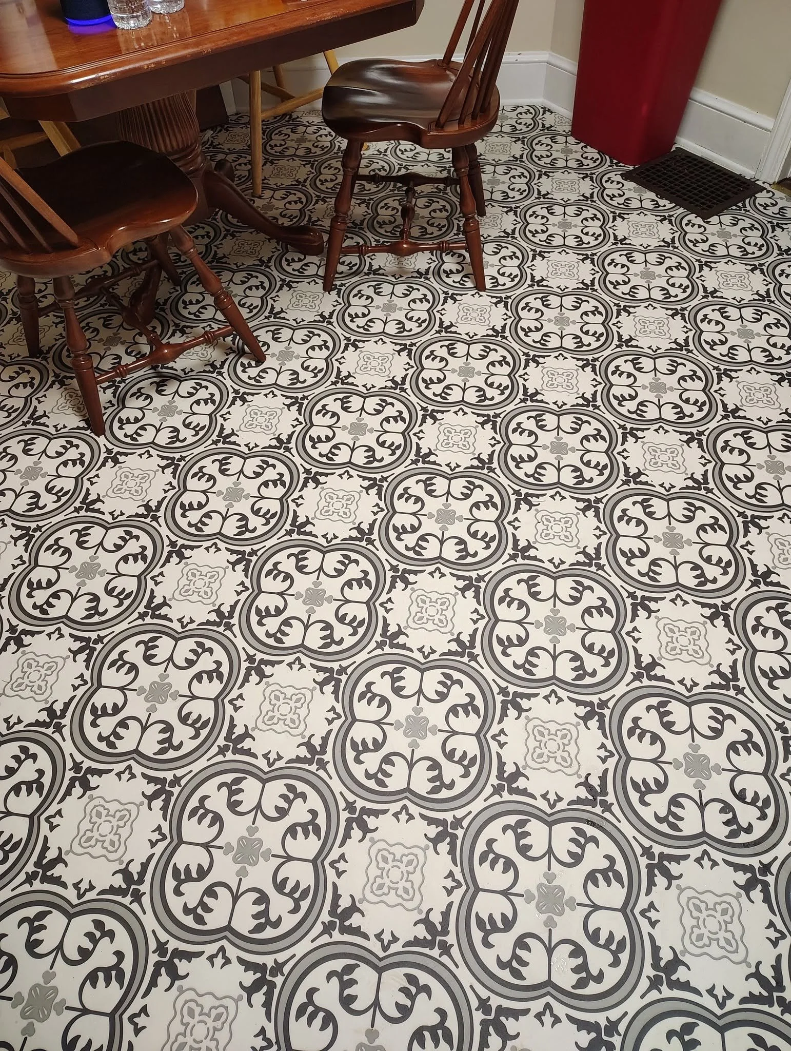 Decorative patterned black and white tile floor in a room with wooden chairs around a table, a red trash can, and a black floor vent.