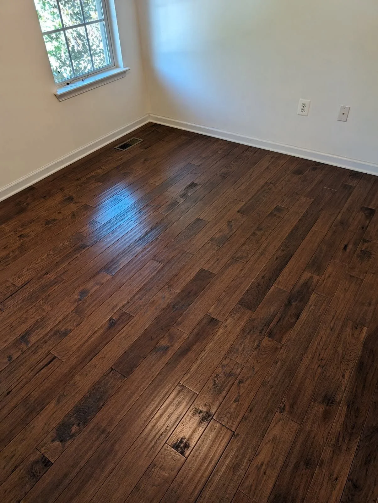 Empty room with dark hardwood flooring, beige walls, a window with natural light, and electrical outlets.