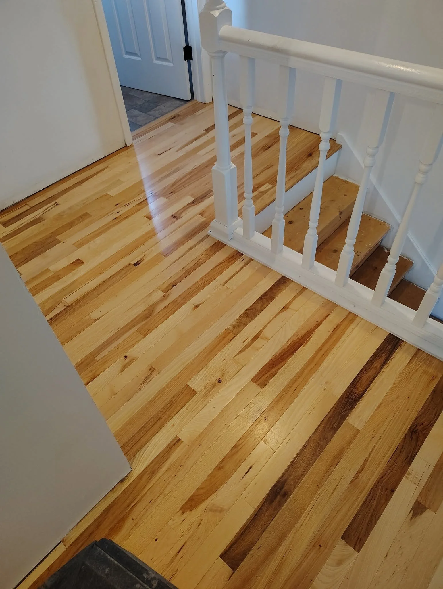 View of a wooden staircase with white balusters and handrail, adjacent to a door, with a wood floor and a tiled area in the background.
