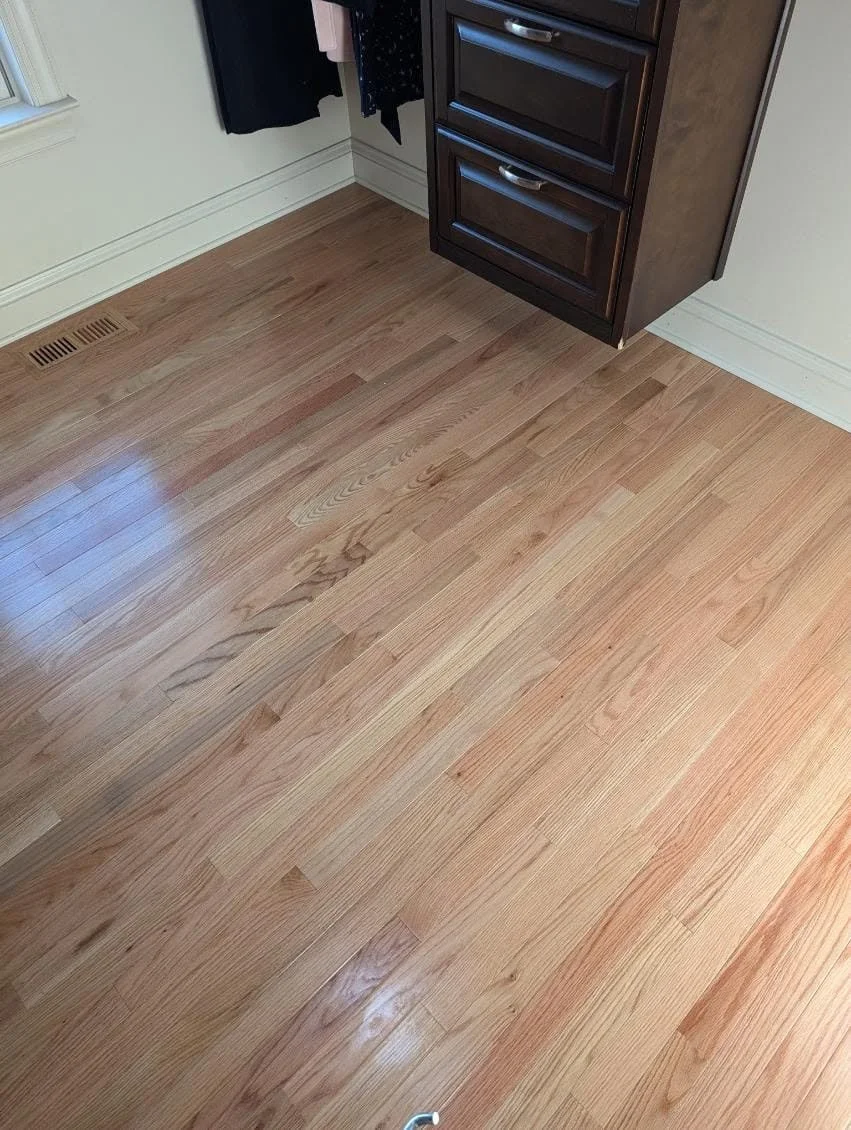 Close-up of a hardwood floor in a room with a dark wooden cabinet and vent on the wall.