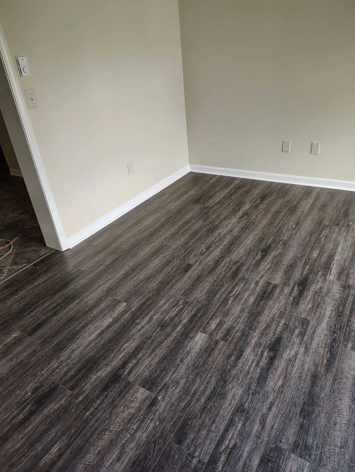 A corner of a room with dark wood laminate flooring, cream walls, and white baseboards. There are electrical outlets and a thermostat on the wall.