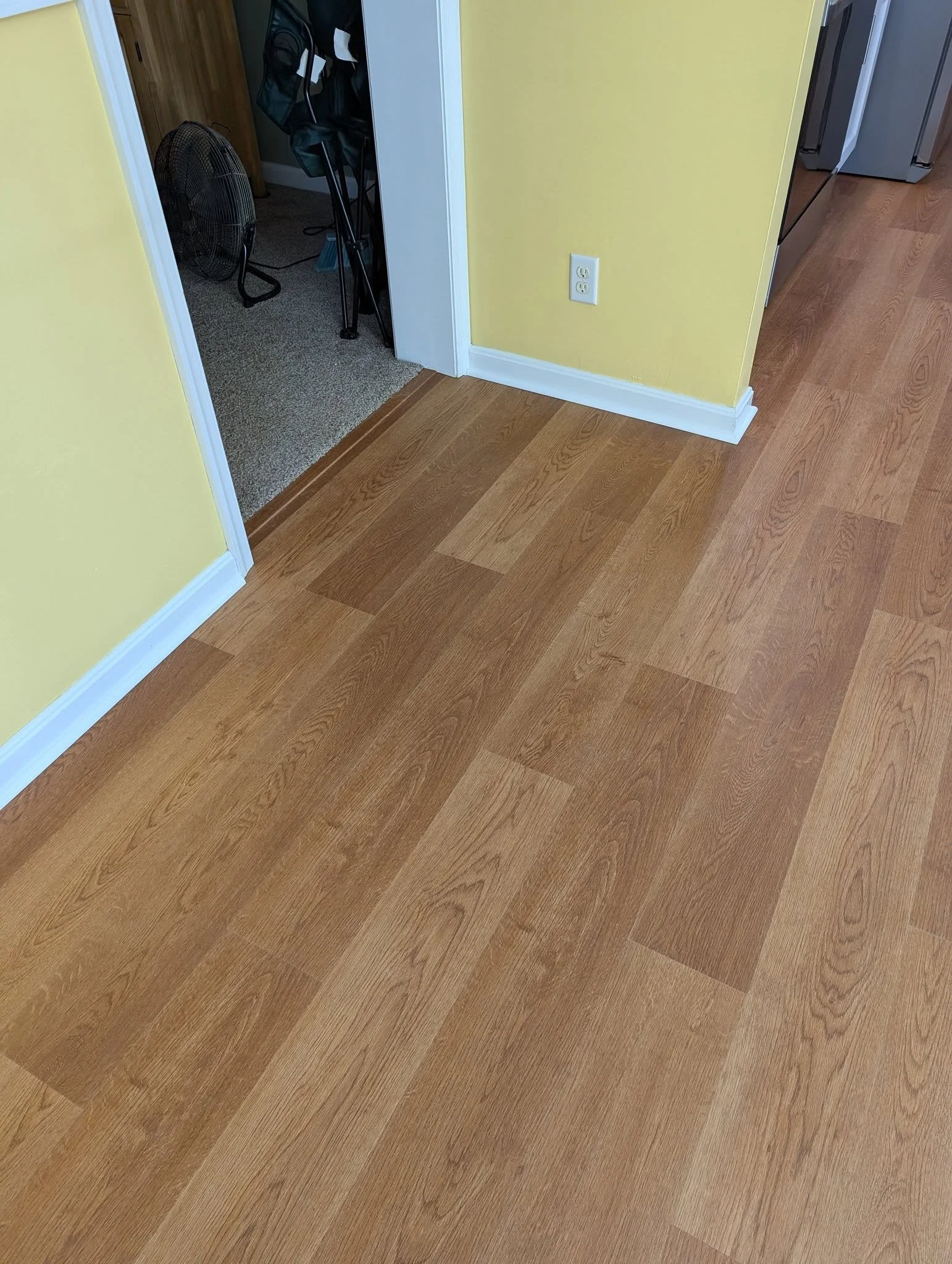 Image of a wooden floor in a room with yellow walls and white baseboards, a doorway leading to a carpeted room with a fan and a table, and a stainless steel refrigerator in the kitchen area.
