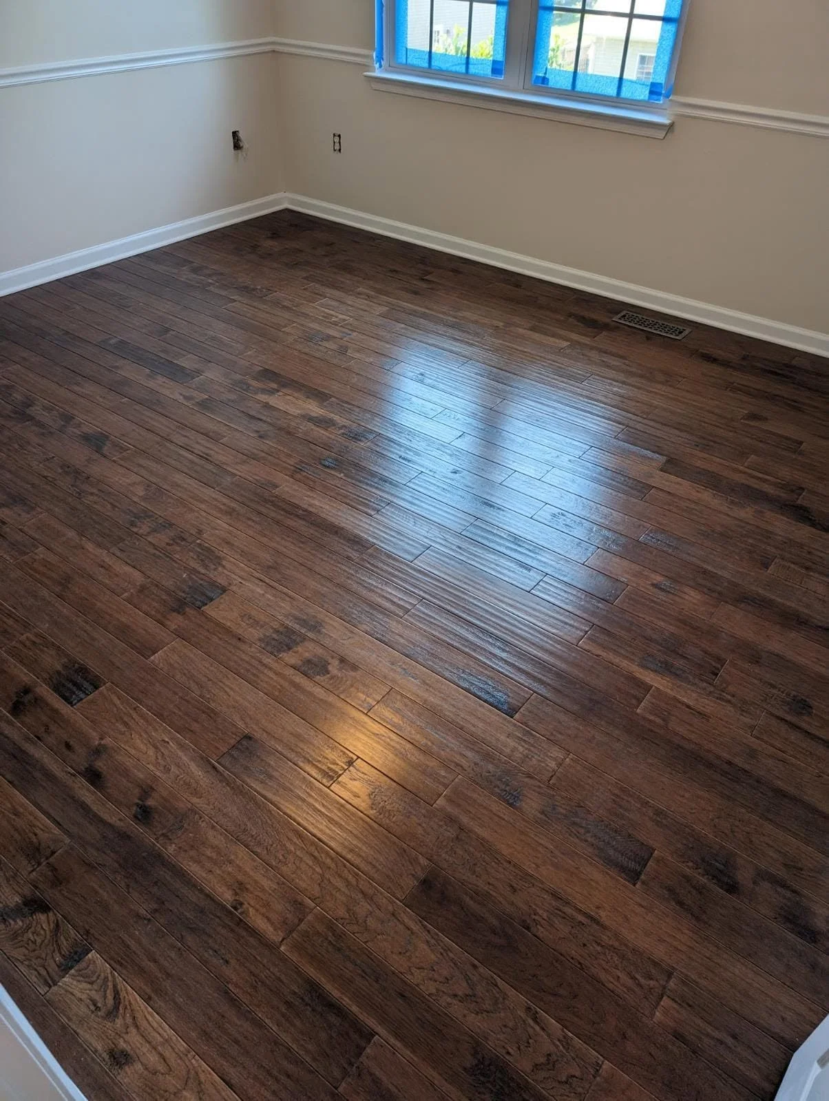 Empty room with wooden flooring, white walls, a window with blue tape, and baseboard heating vent.