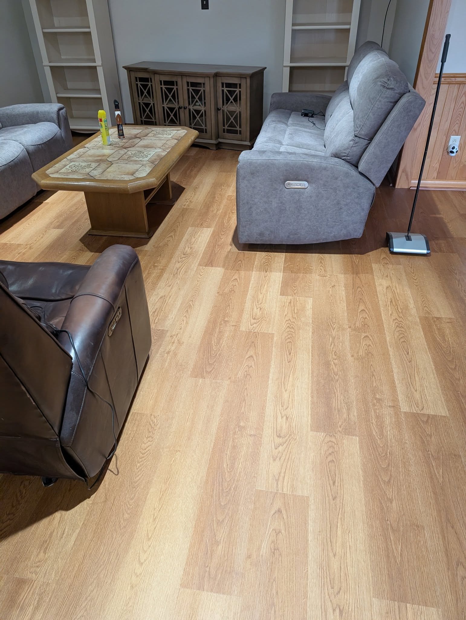 Living room with hardwood floor, gray sofa, brown armchair, coffee table, and shelves in the background.