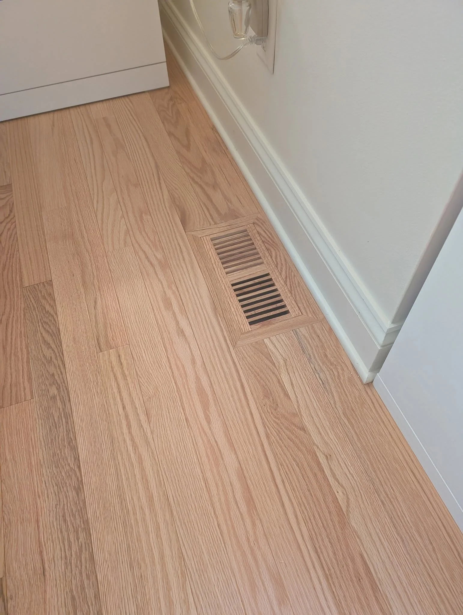 Close-up of a kitchen floor with wood flooring and a metal vent near the bottom right corner, white baseboard along the wall, and an electrical outlet with a plug.