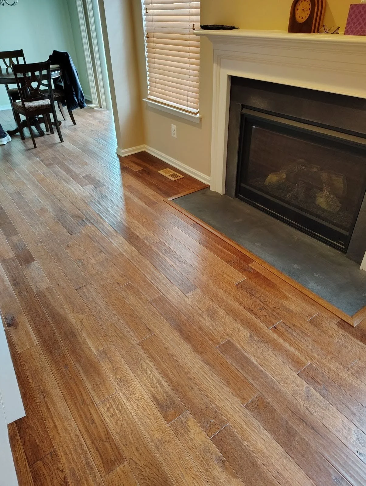 Wooden floor in a living room with a fireplace and window with blinds, part of a dining area with chairs and table in the background.