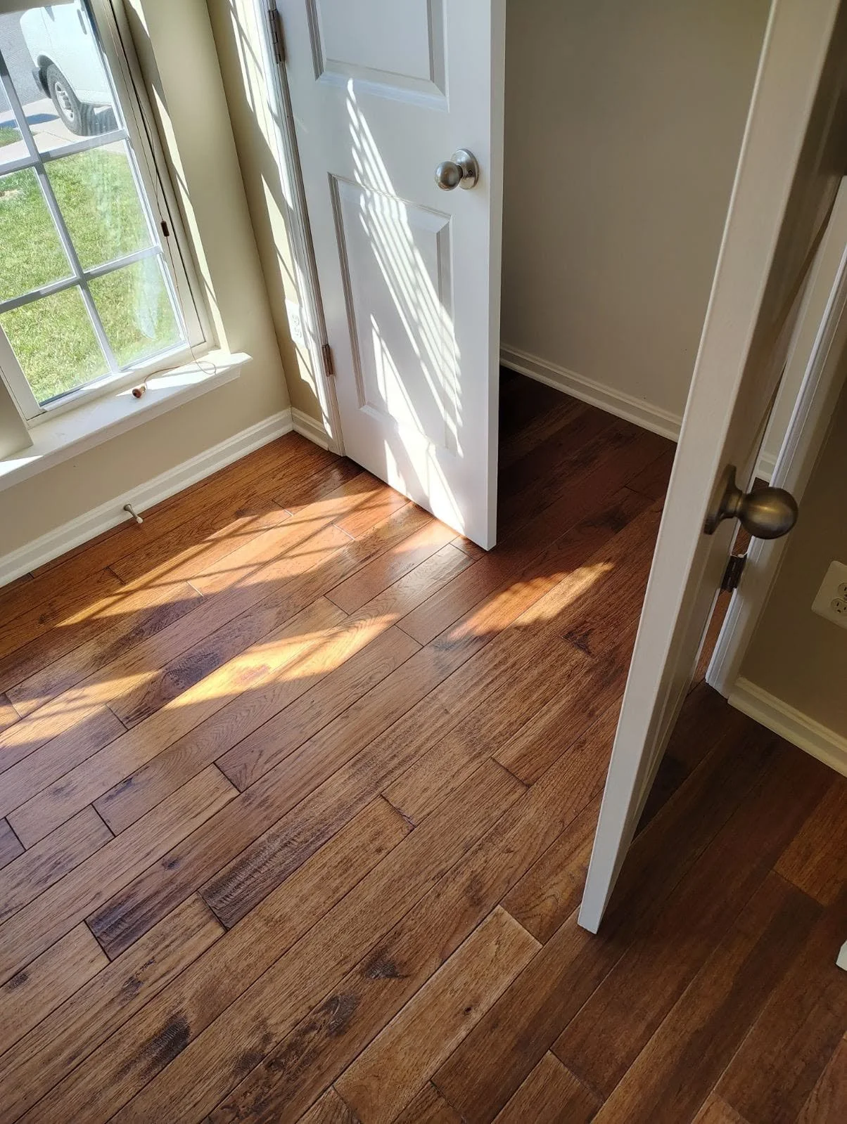 Sunlight casting shadows on a wooden floor inside a house, with an open closet door and a window showing a grassy yard and parked vehicle outside.