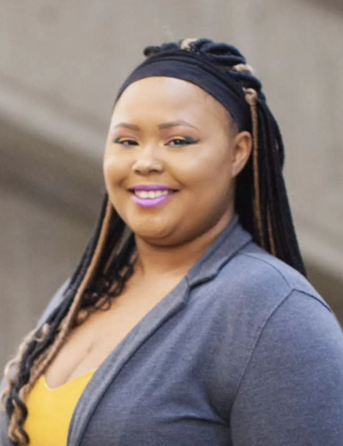 A woman smiling, with long braided hair, wearing a gray blazer and yellow top, against a blurred background.
