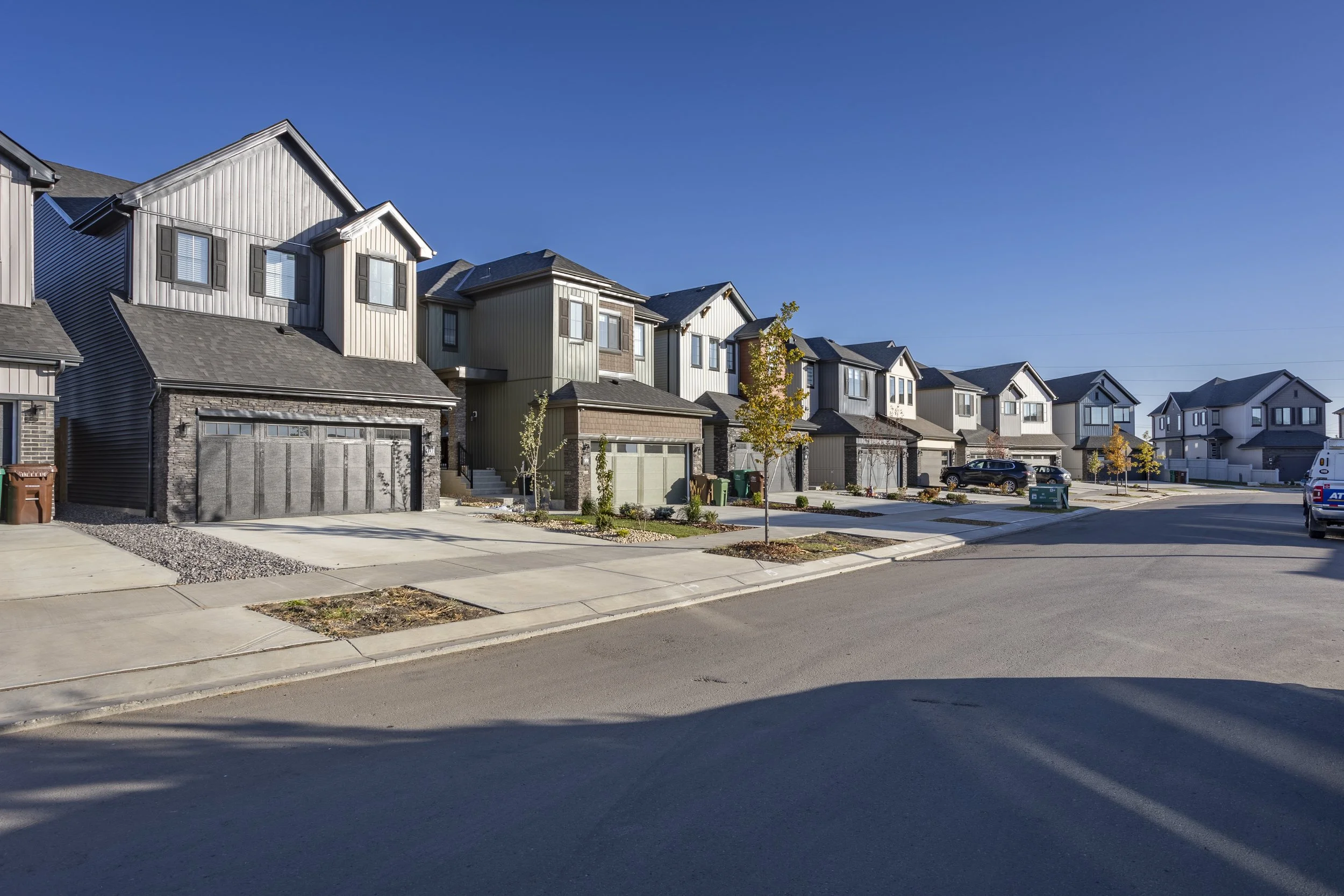 A row of modern suburban houses with garages, young trees, and parked cars on a sunny day.