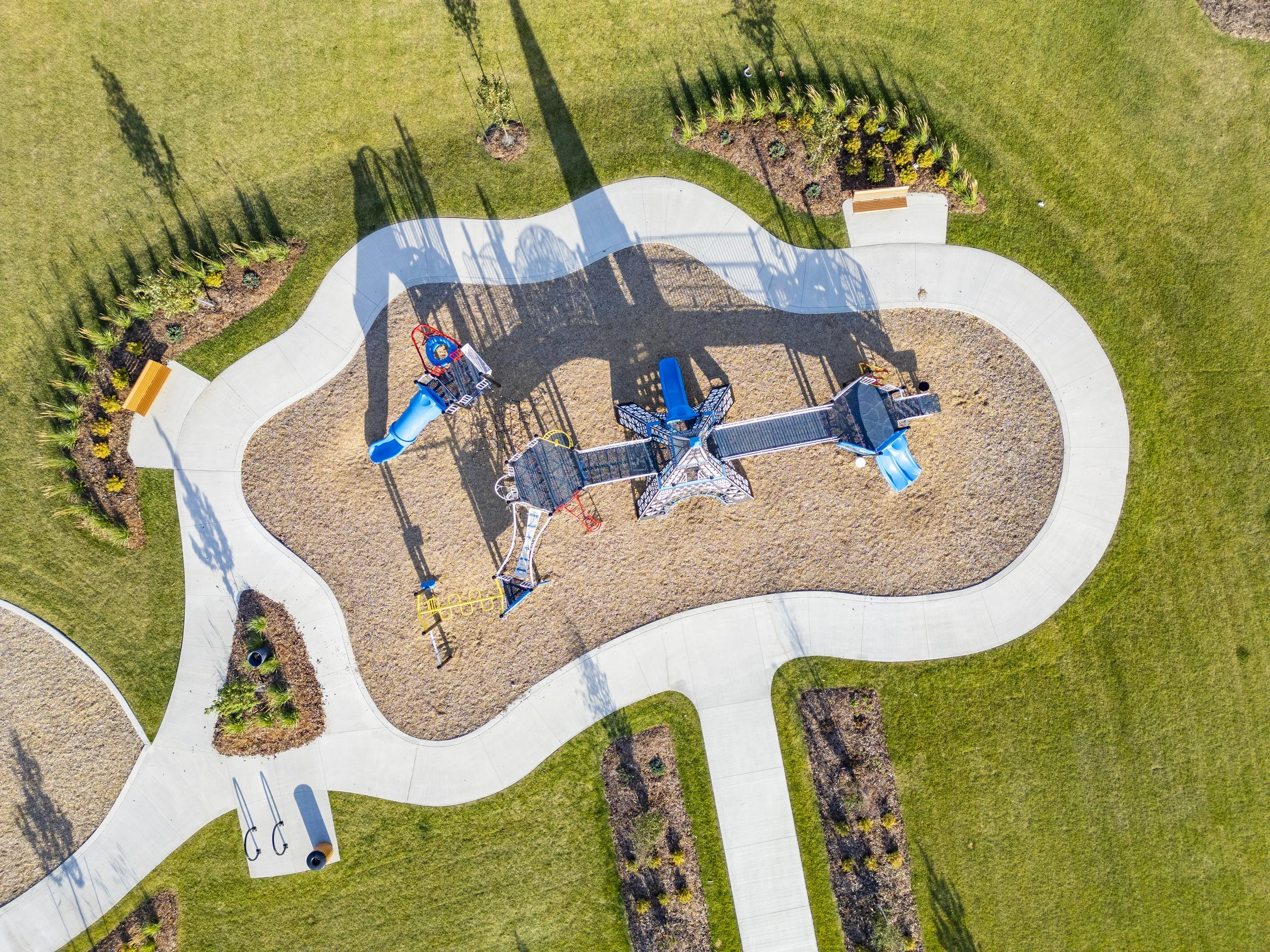 An aerial view of a playground with a winding paved path surrounding it, green grass, landscaped flower beds with plants, a bench, and a children's slide set with shadows cast by nearby trees.