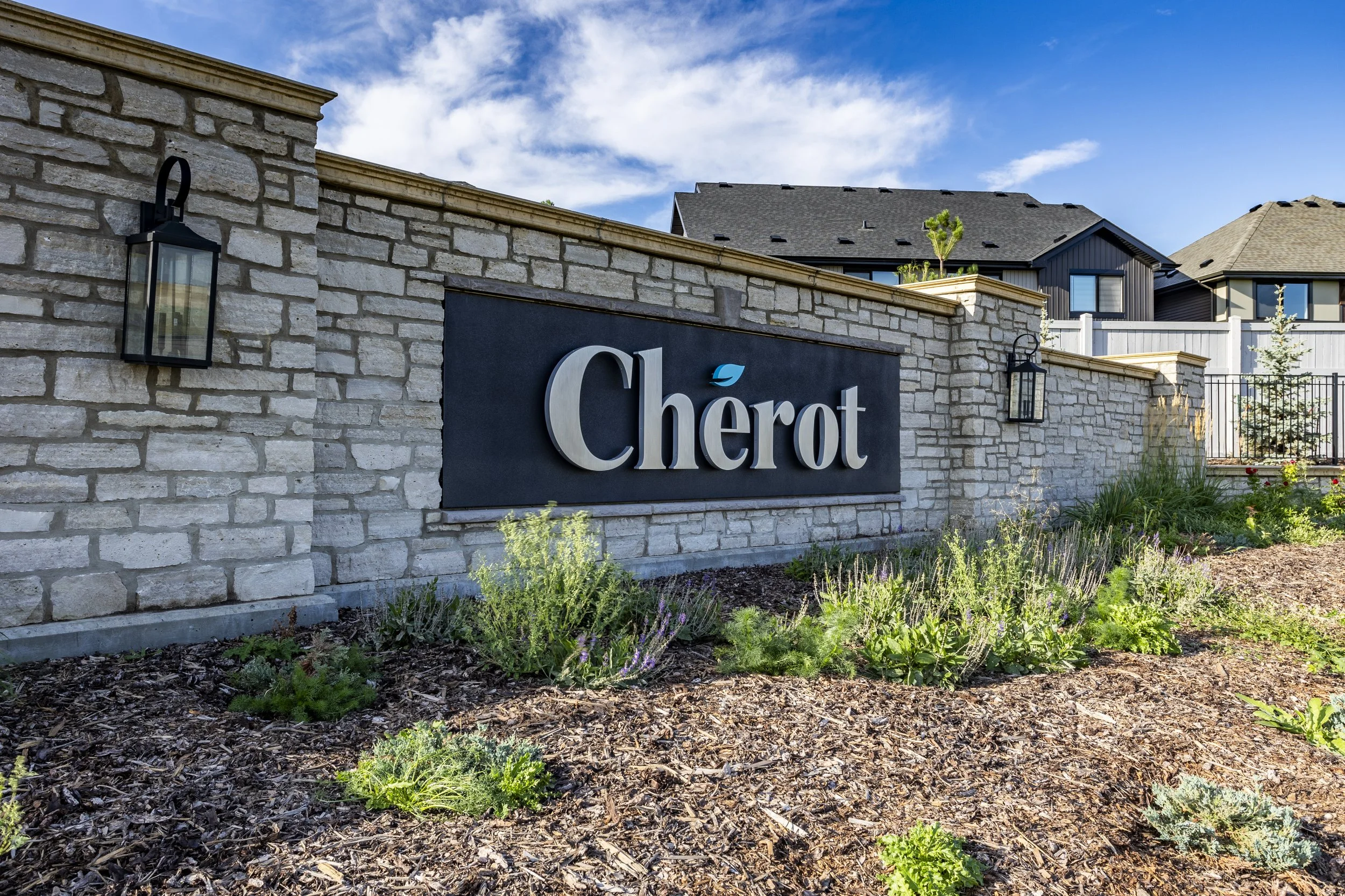 A stone wall with a black sign titled 'Chérôt' and two black lanterns on either side. Behind the wall, there are rooftops of modern houses, some trees, a clear blue sky with scattered clouds, and landscaped plants and flowers in the foreground.