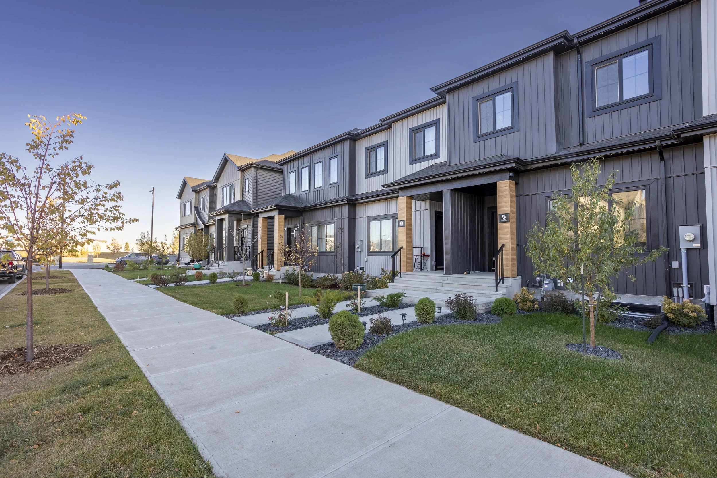 A row of modern townhouses with front porches and small steps, landscaped front yards with young trees and shrubs, along a concrete sidewalk under a clear blue sky.