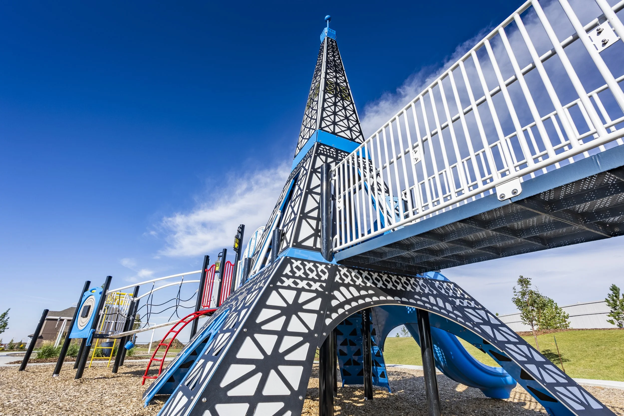 A modern blue and black playground structure with a slide, climbing features, and a tower, set outdoors under a blue sky with clouds.