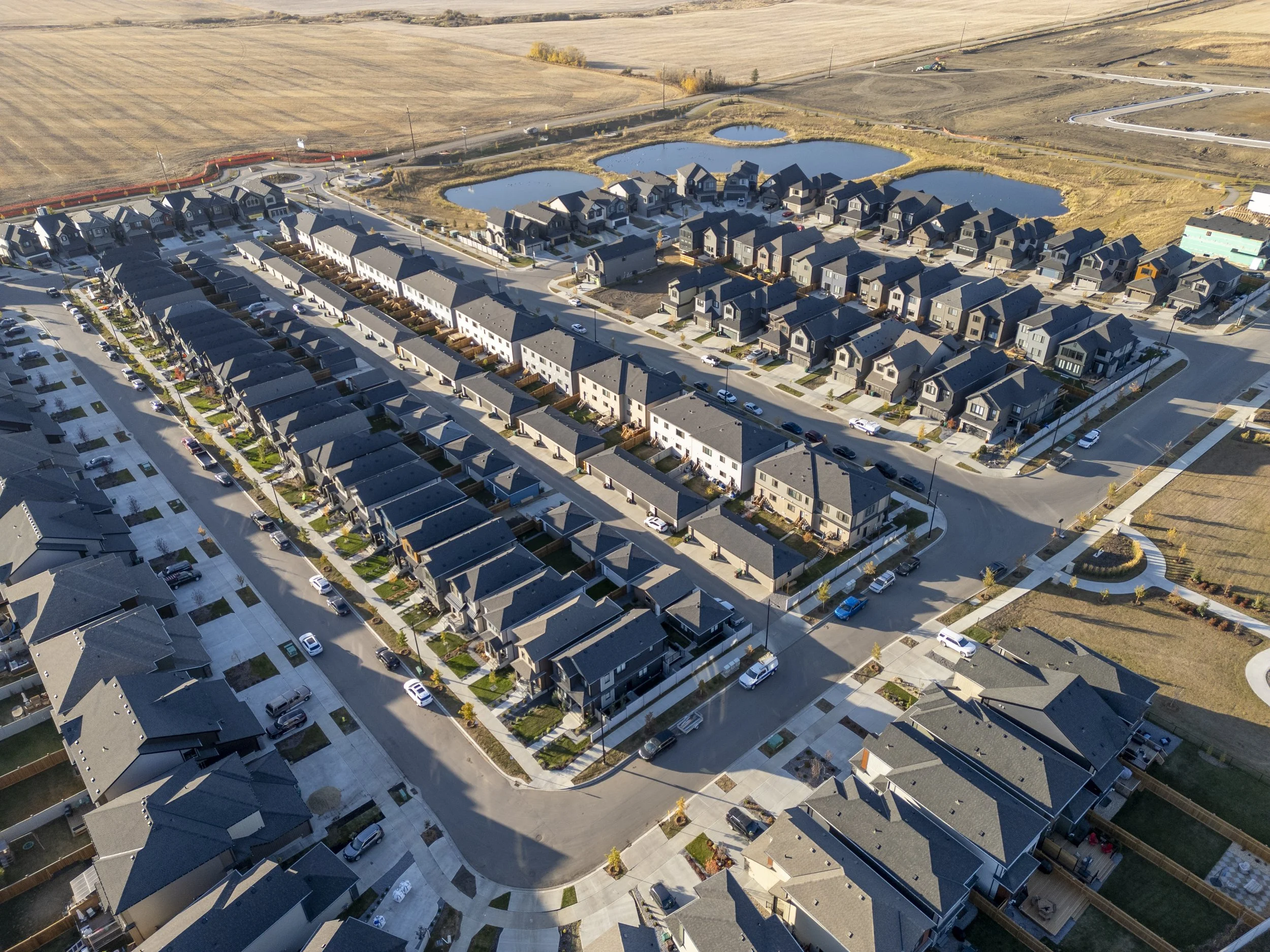 An aerial view of a residential neighborhood with rows of houses, ponds, and streets. The area is surrounded by open fields and farmland.