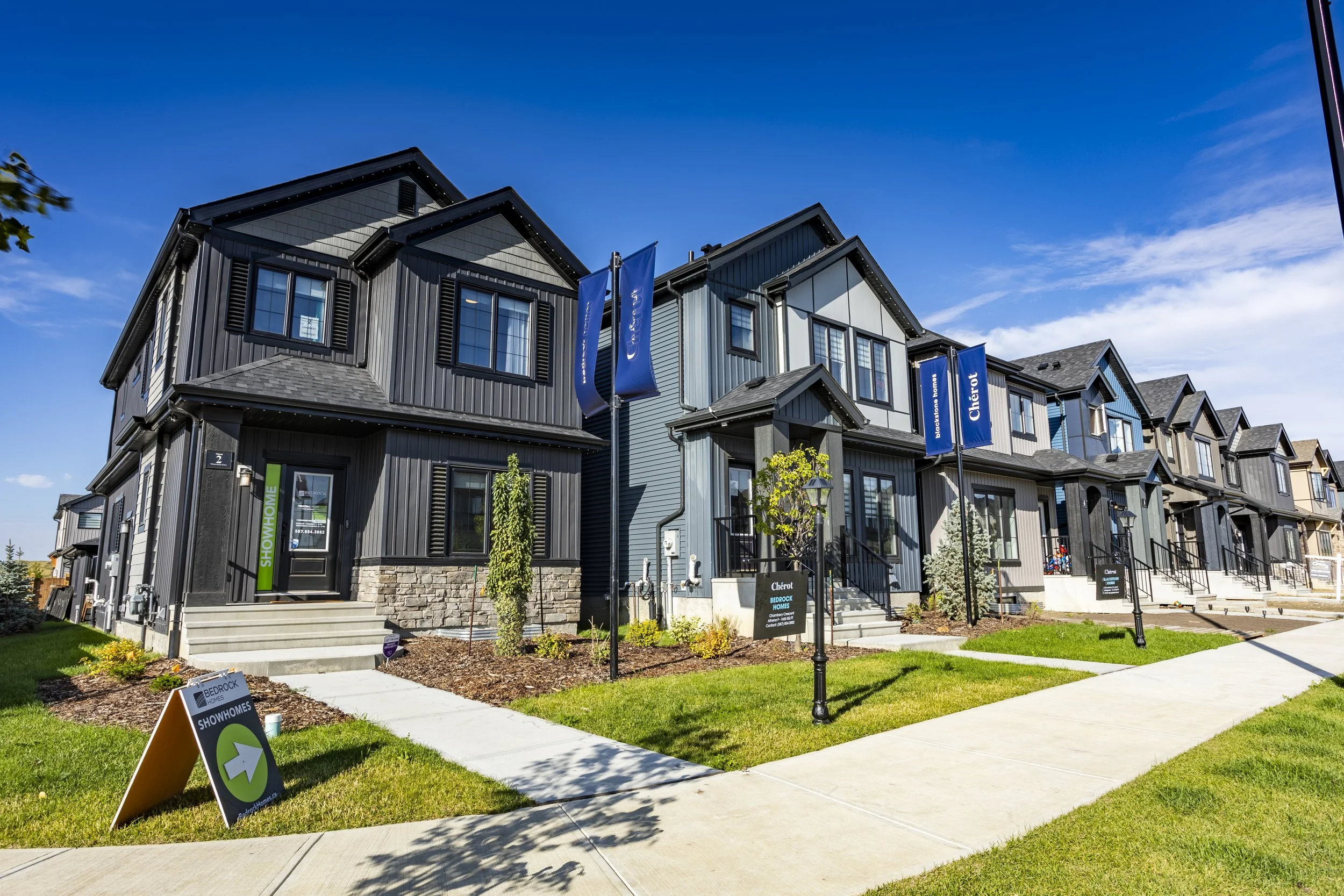 A row of modern townhouses with dark gray and light gray siding, black shutters, and small front gardens. Blue flags and signs indicate the homes are available for showings or sale. The sky is clear with some wispy clouds.