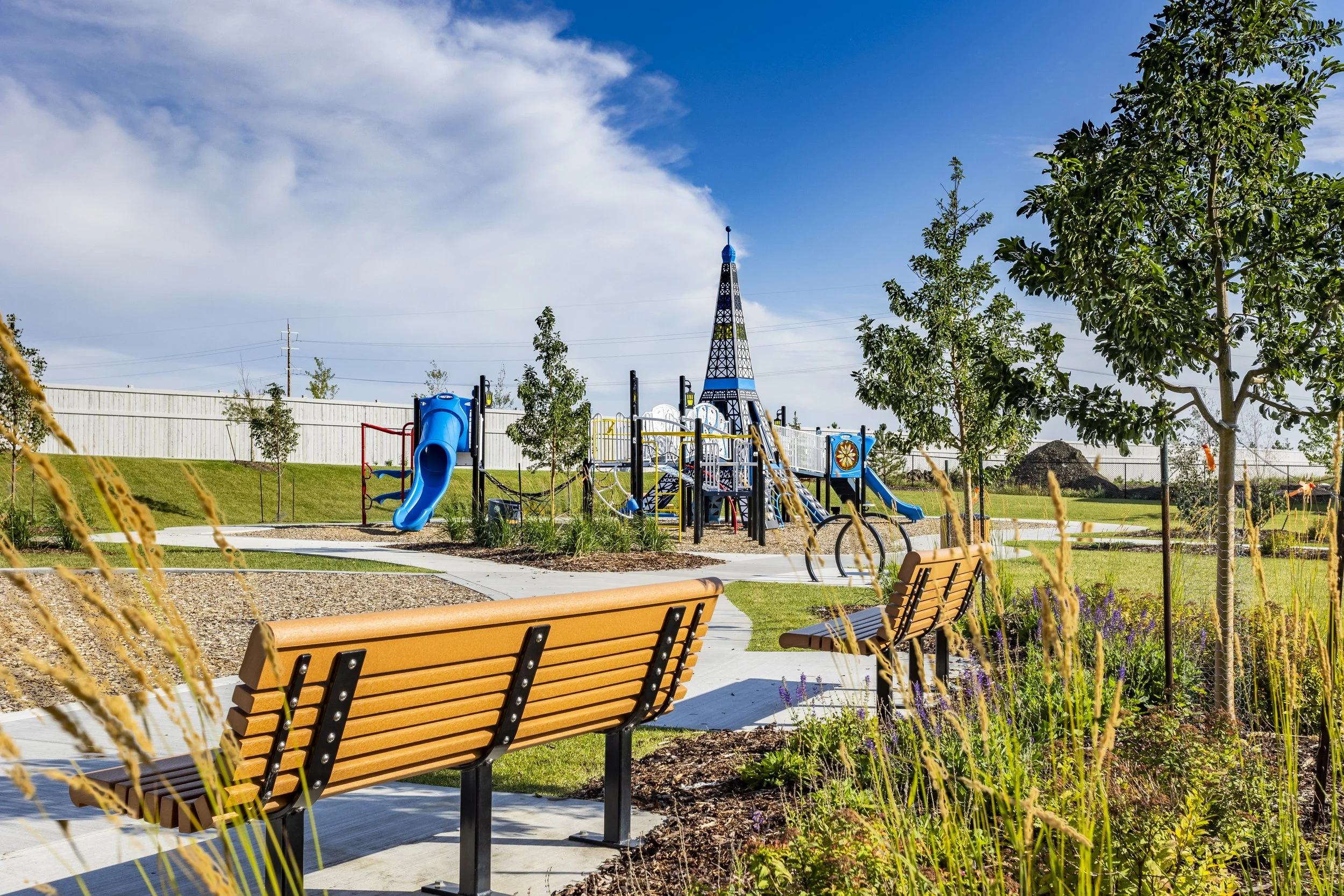 Playground with slides, climbing structures, and a tall tower in a park, with trees, benches, and a clear blue sky.