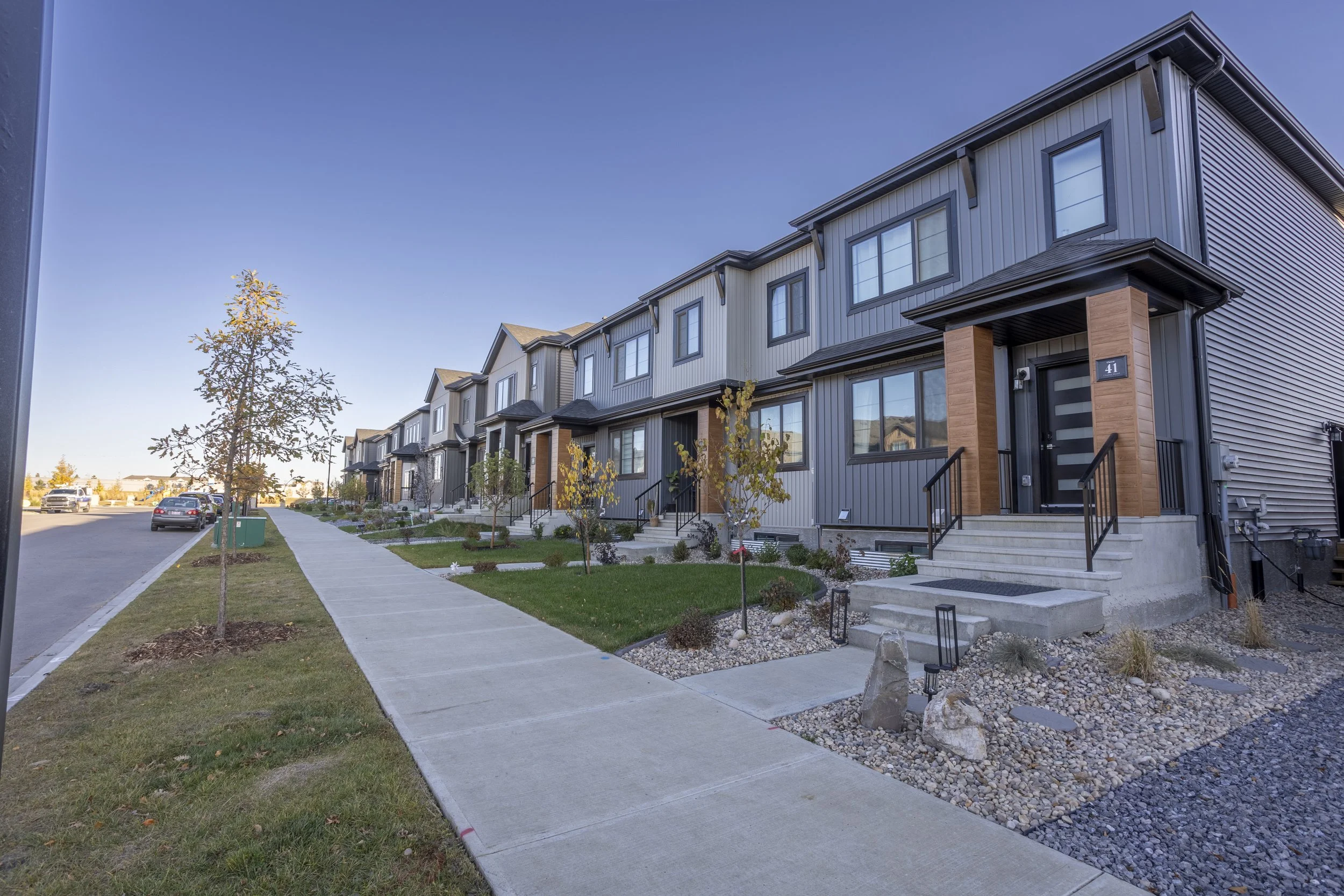 A row of modern townhouses with metal siding, black trim, and small front steps, along a concrete sidewalk and street with parked cars, under a clear blue sky with a few trees.