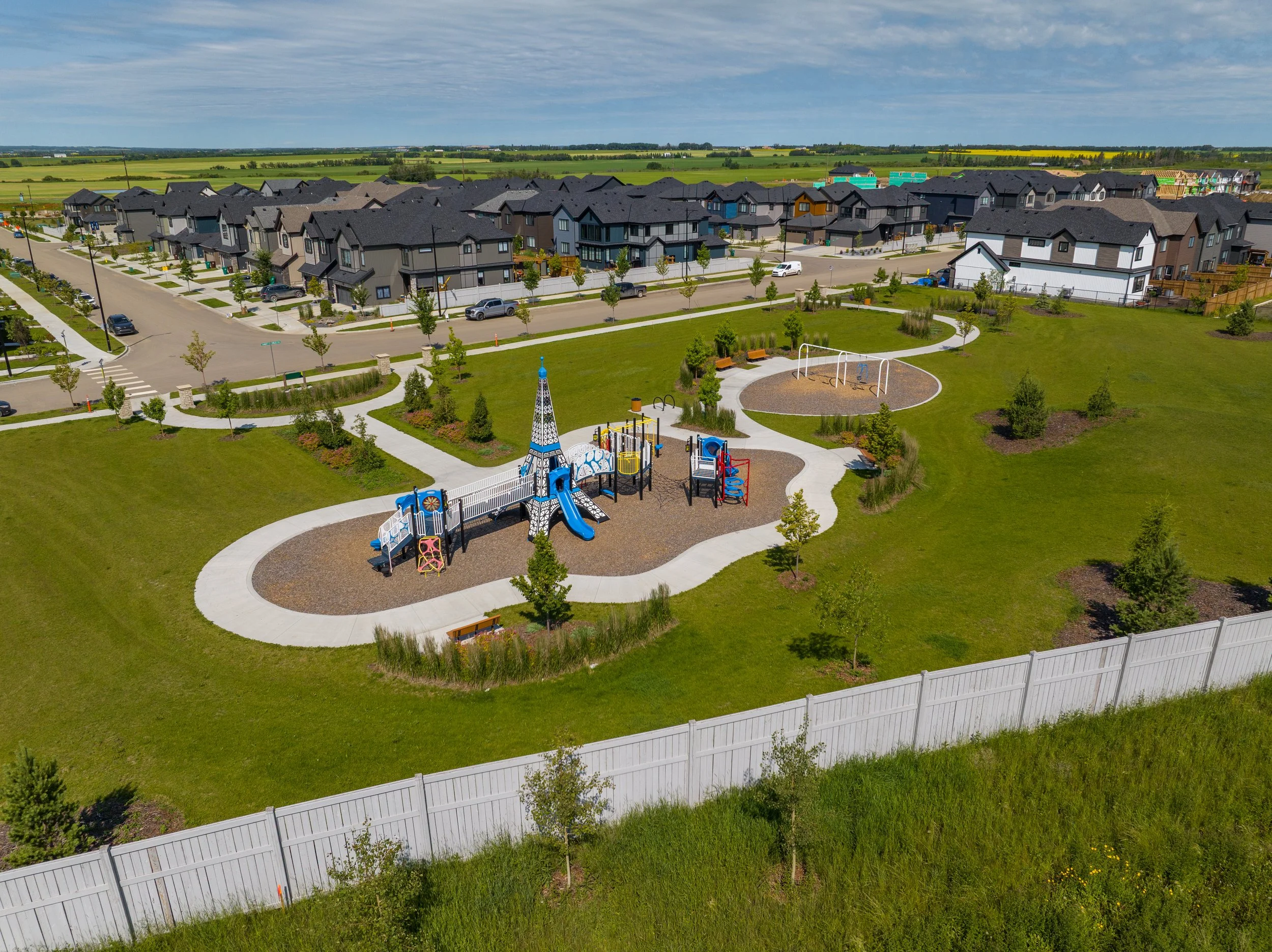 Aerial view of a suburban neighborhood with modern houses, a park area with playground equipment and picnic tables, and open fields in the background under a partly cloudy sky.