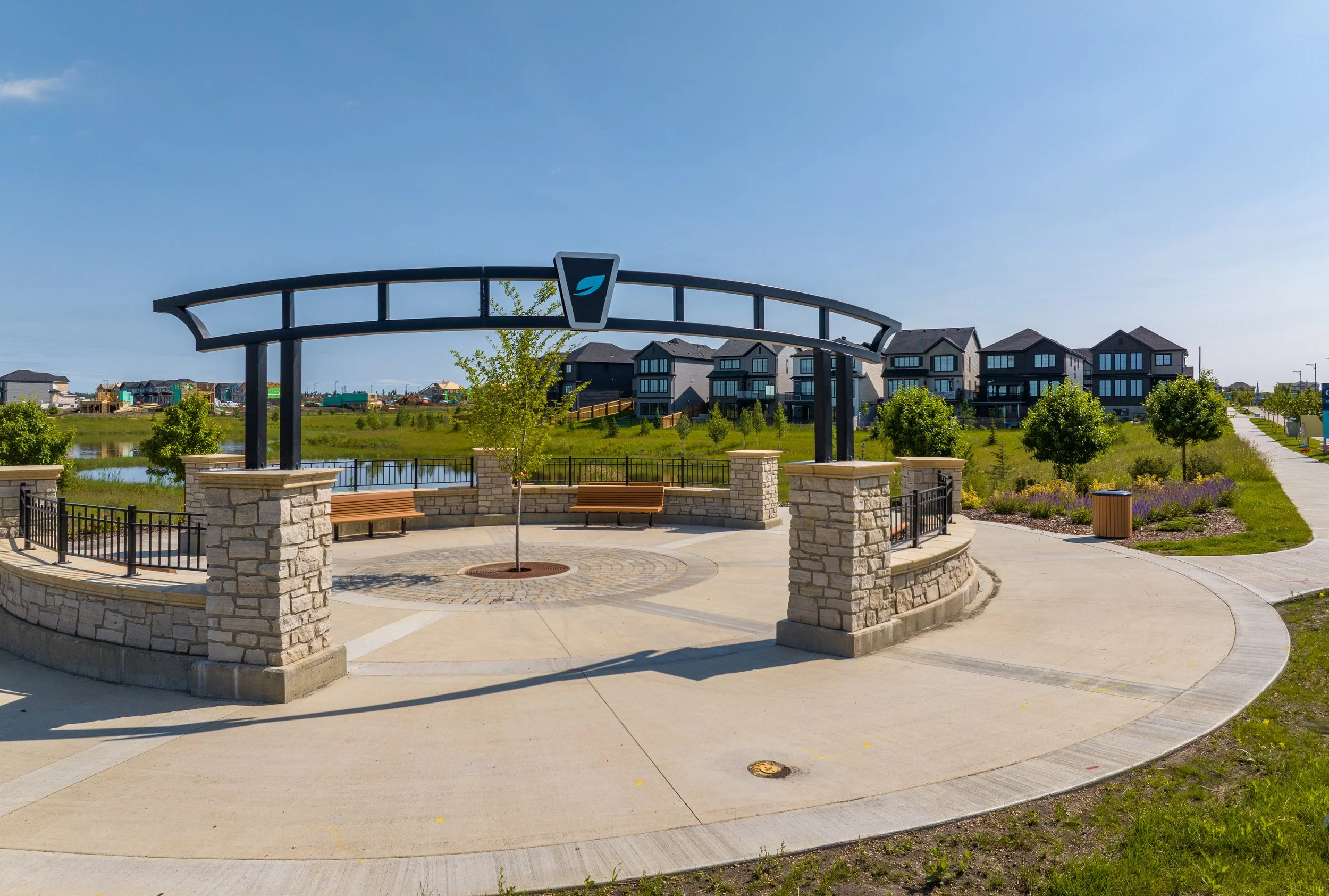 A park with a circular stone and concrete seating area, surrounded by a black metal fence, trees, and a walking path. In the background, there are modern houses and a body of water. A decorative metal arch with a logo is at the entrance of the seating area.
