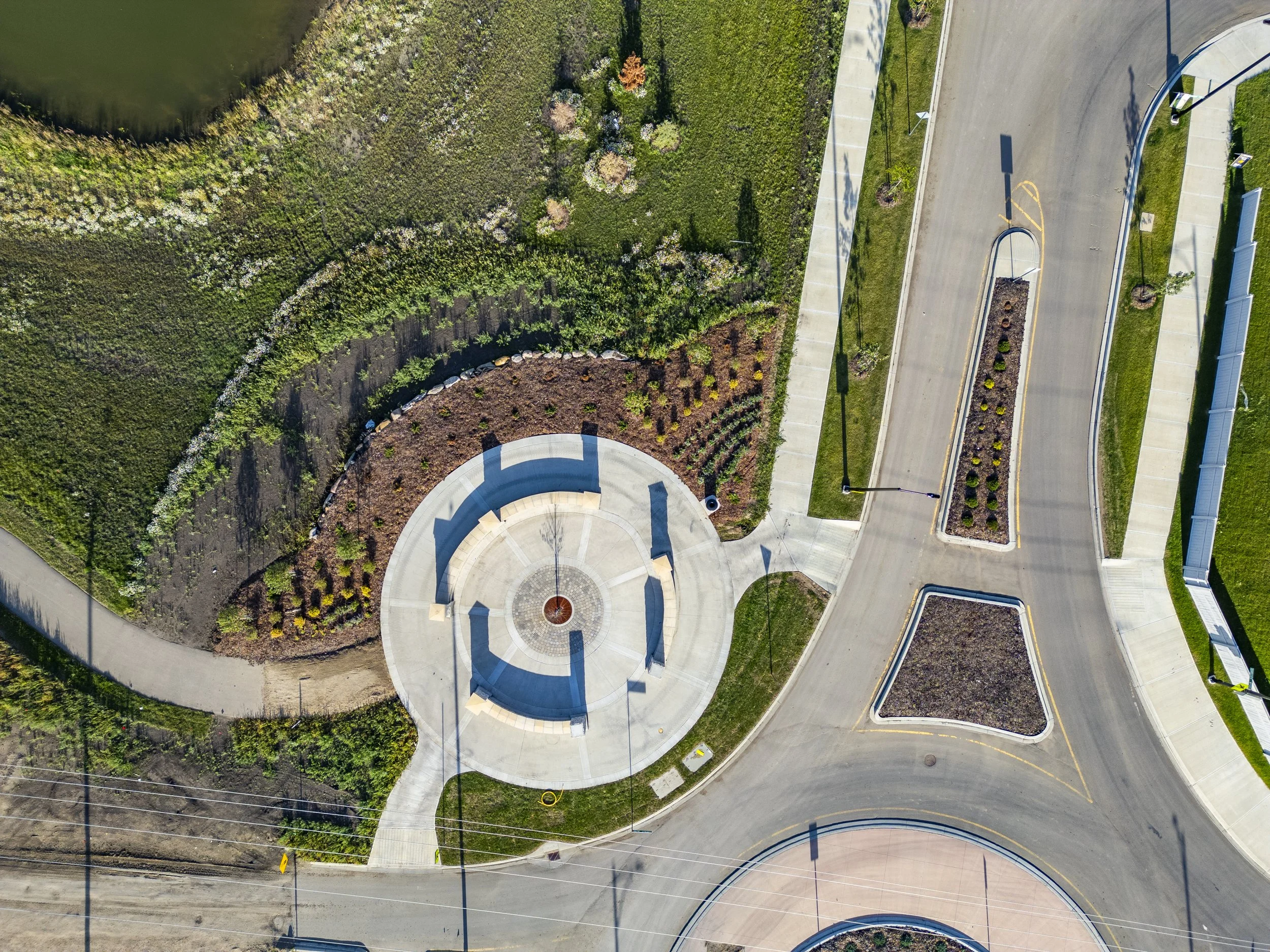 An aerial view of a park with curved pathways, green grass, and landscaped areas, including a small pond on the top left and a circular paved area in the center with a fountain.