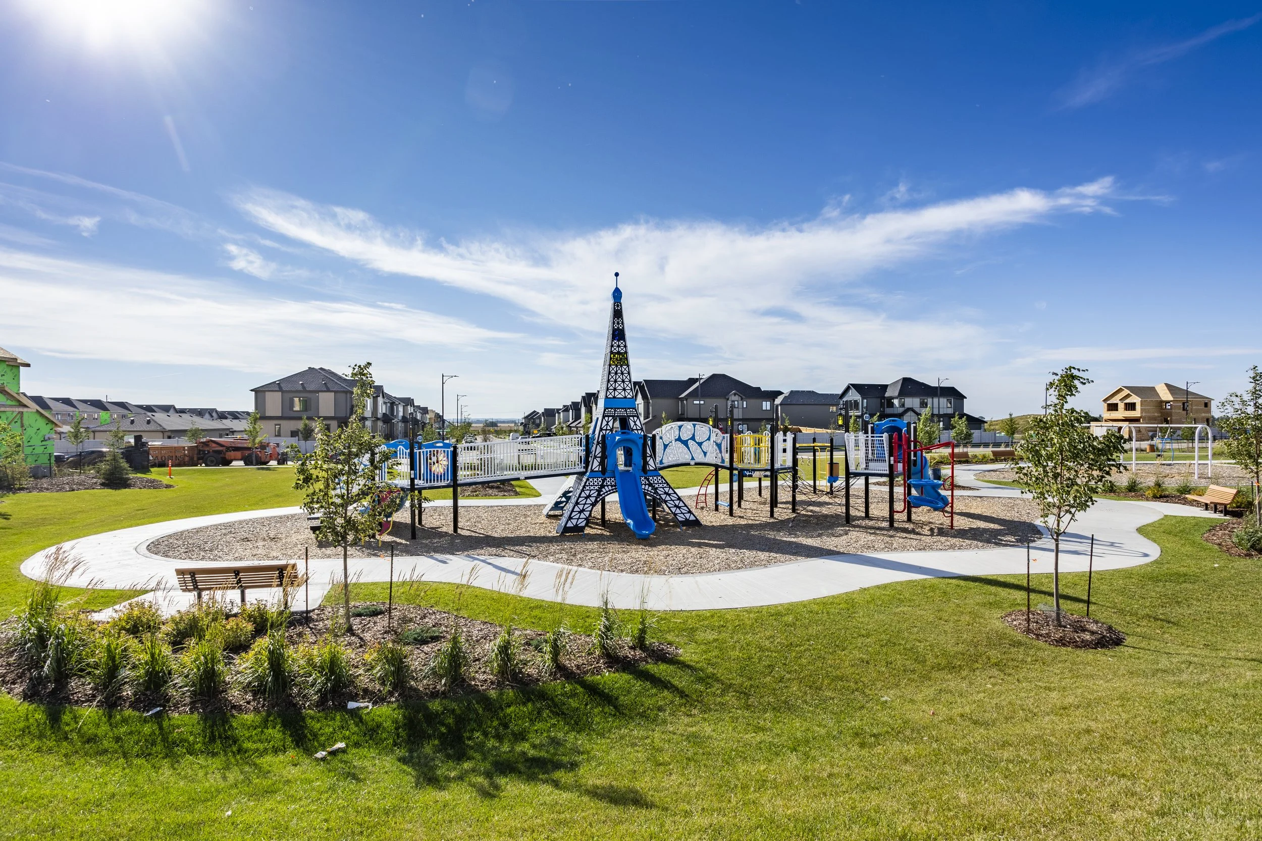 A playground with a large Eiffel Tower-shaped slide, surrounded by a paved walking path, benches, trees, and residential houses under a bright sunny sky.