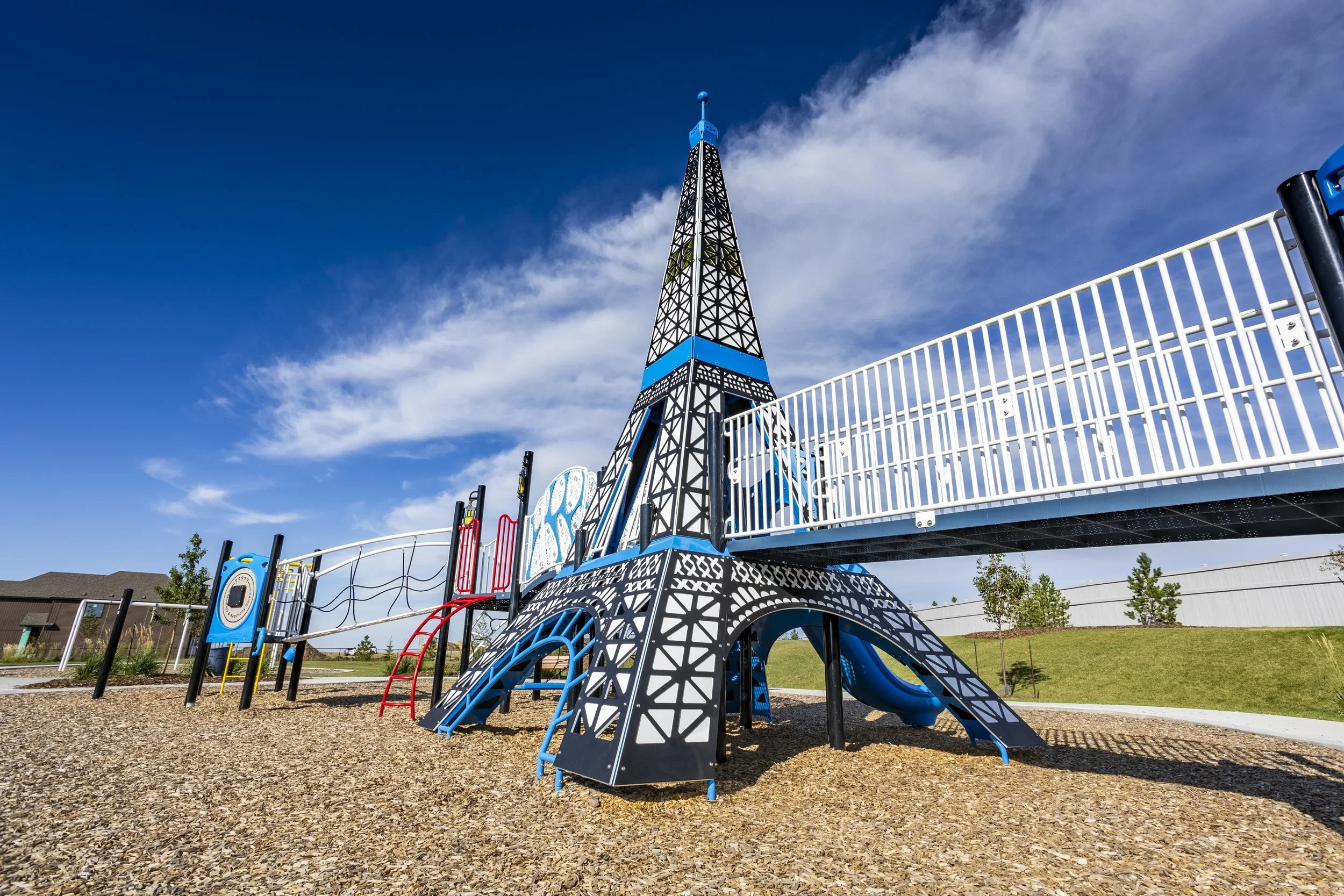 A playground structure designed to resemble the Eiffel Tower, painted in black, blue, and white, with slides and climbing features, set on a wood-chip ground under a blue sky with scattered clouds.