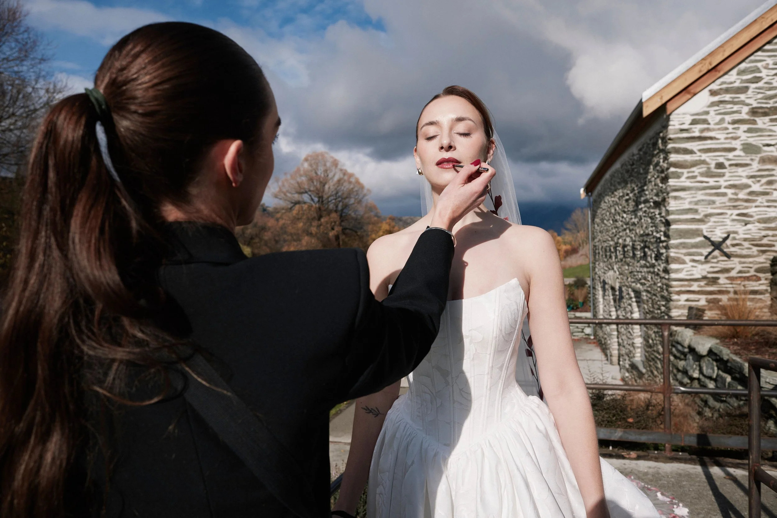 A woman with white skin and red lipstick is getting her makeup done by another woman with red hair tied in a ponytail. The setting appears to be outdoors near a stone building with slate roof, trees, and cloudy sky.