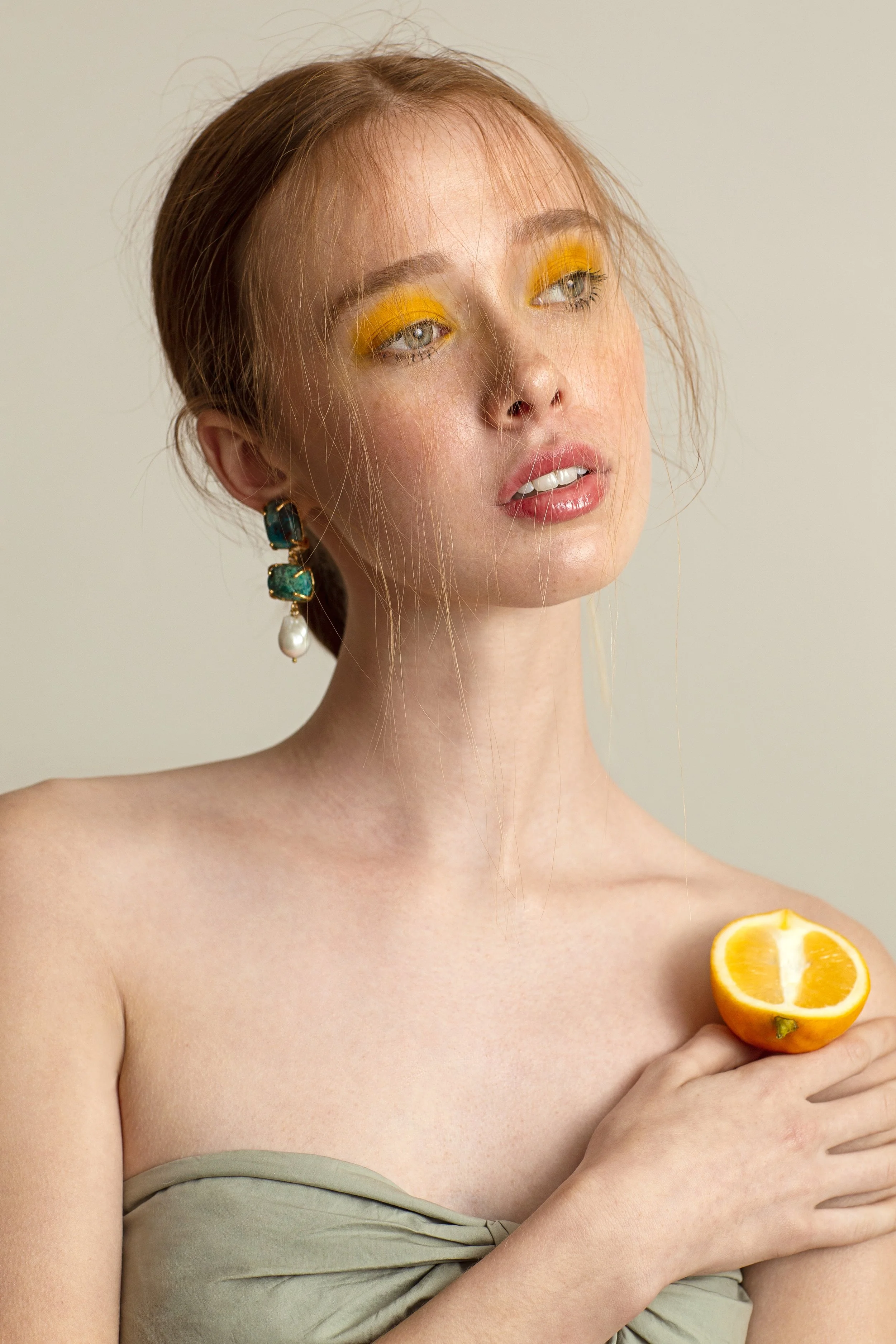 A woman with red hair, blue eyes, and wearing bright yellow eyeshadow is holding a halved orange in her hand, with an additional orange half visible on her shoulder. She wears large earrings with blue stones and a pearl, and is dressed in a light green strapless top.
