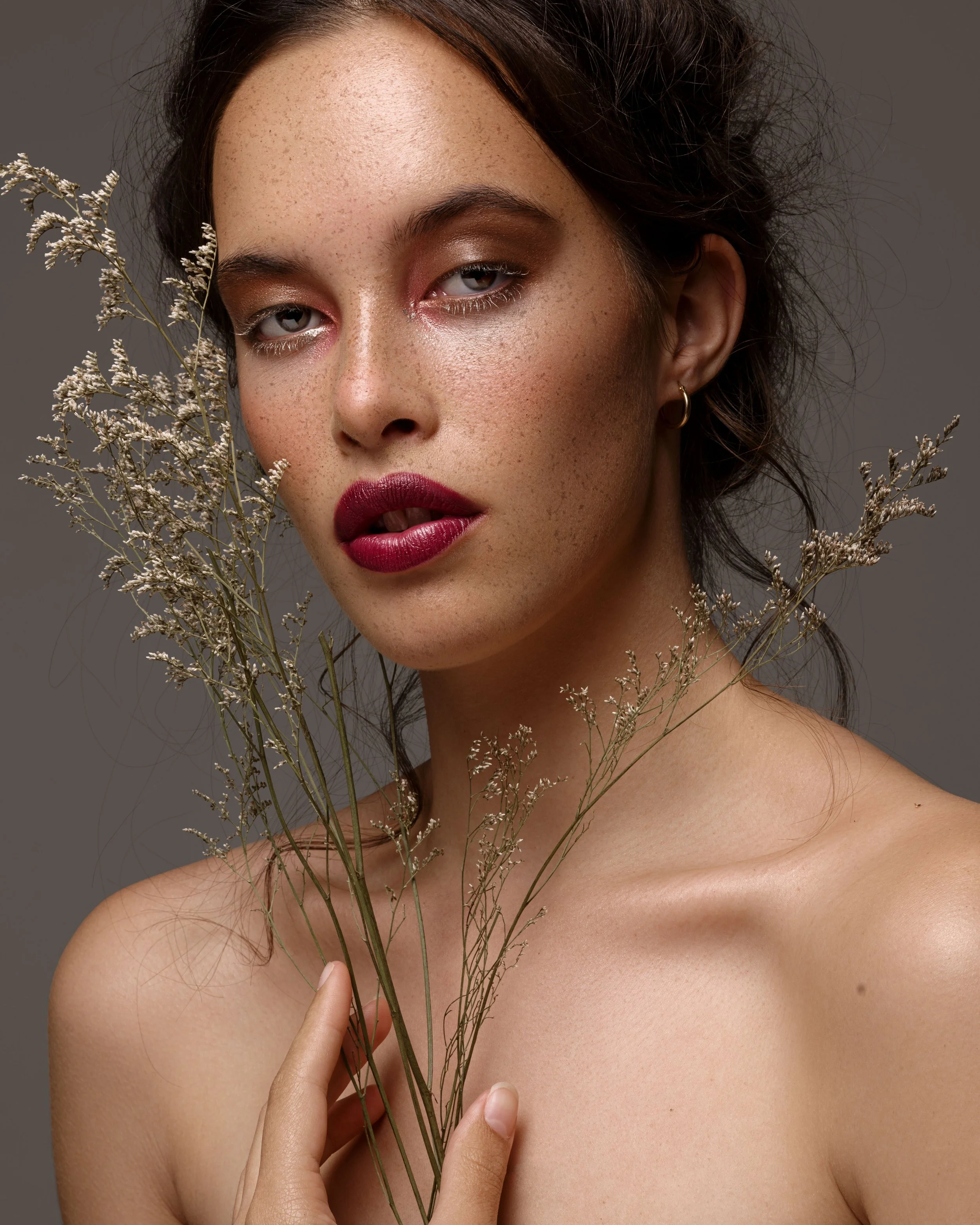 Close-up of a woman holding dried flowers near her face, with makeup emphasizing her eyes and lips, and a neutral background.