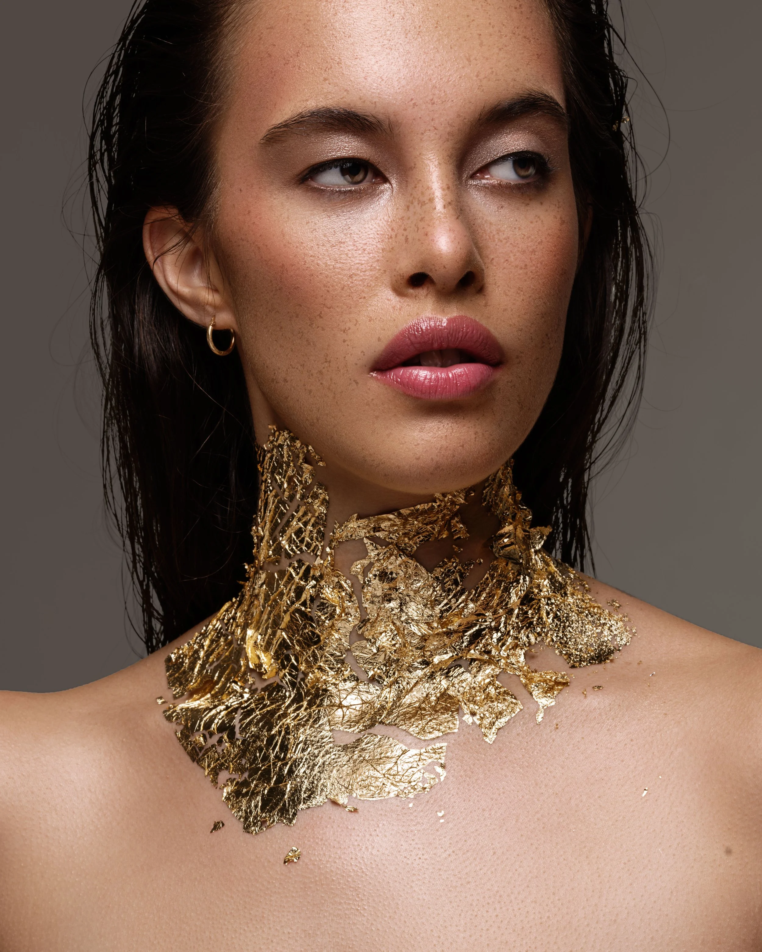 Close-up of a woman with tan skin and freckles, wearing gold jewelry, with gold foil on her chest and neck.