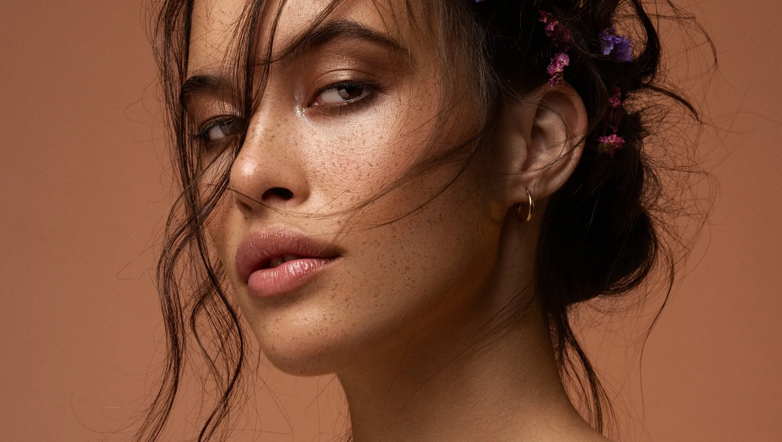 A close-up portrait of a woman with dark hair, freckles, and natural makeup, wearing a small hoop earring and a flower hair accessory, against a neutral background.
