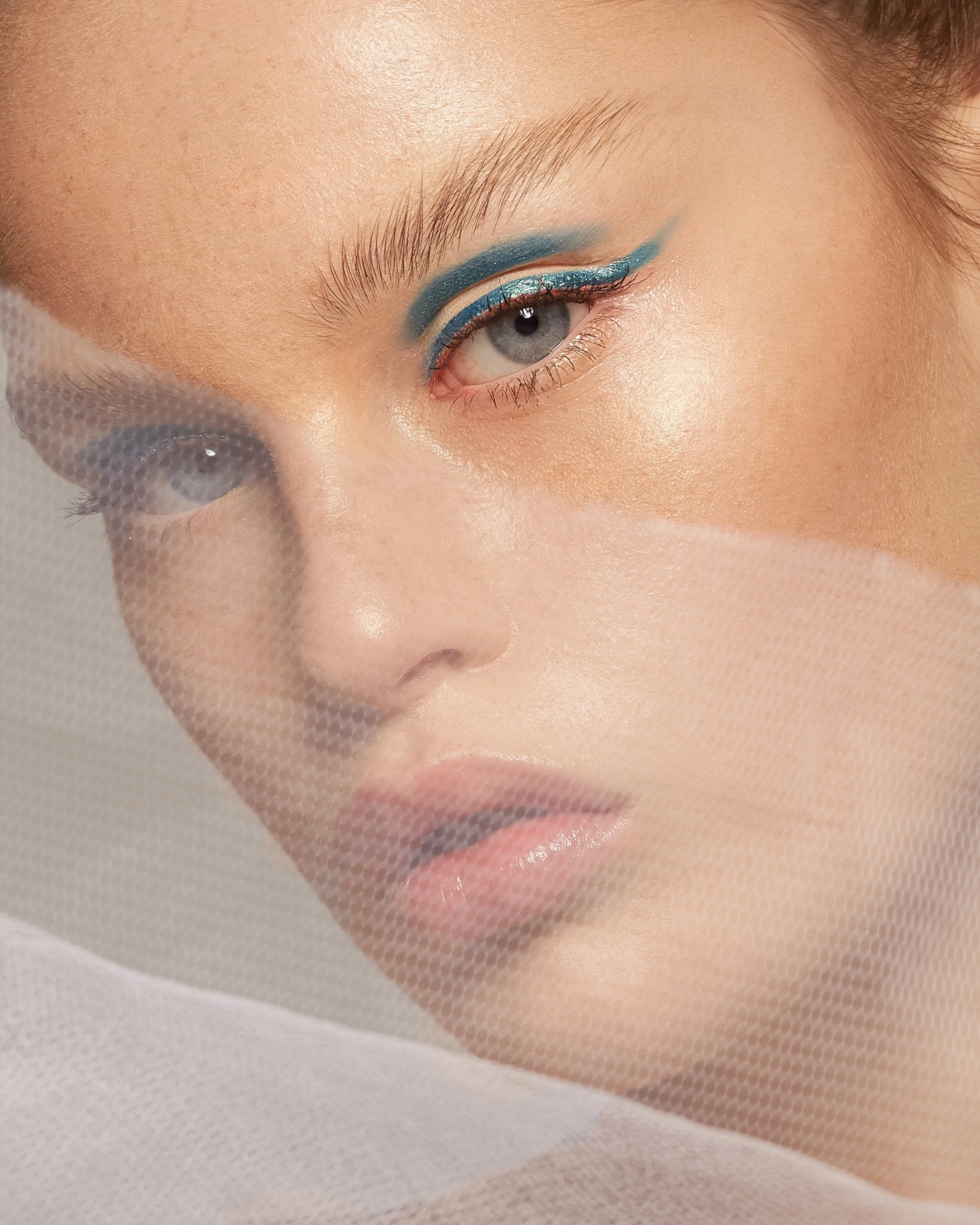 Close-up of a woman's face emphasizing her eye with blue eyeshadow and eyeliner, and a netted fabric partially covering her face.