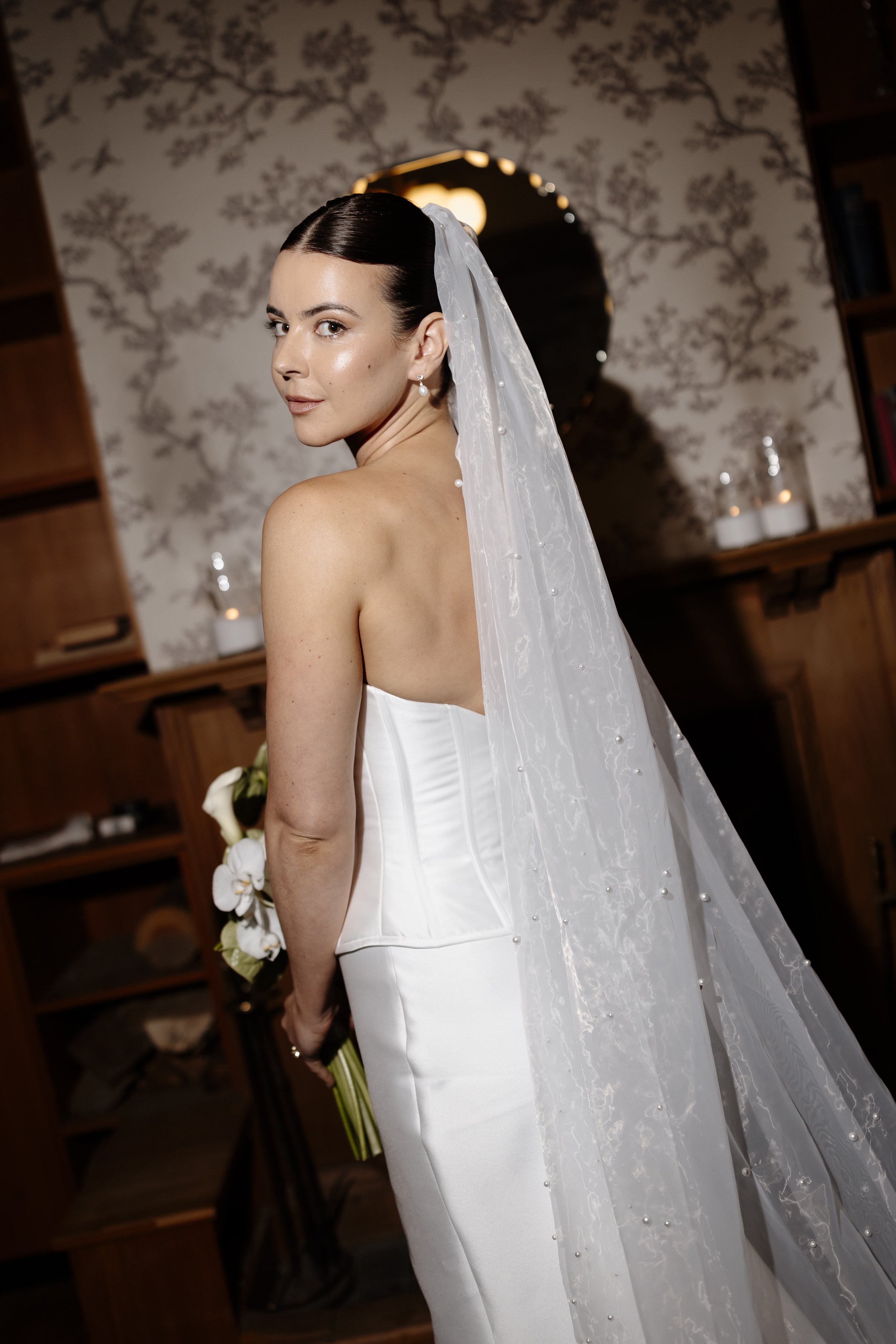 A bride in a white strapless wedding dress with pearl accessories and a veil stands indoors in front of a decorative wall and mirror, holding a bouquet of white flowers.