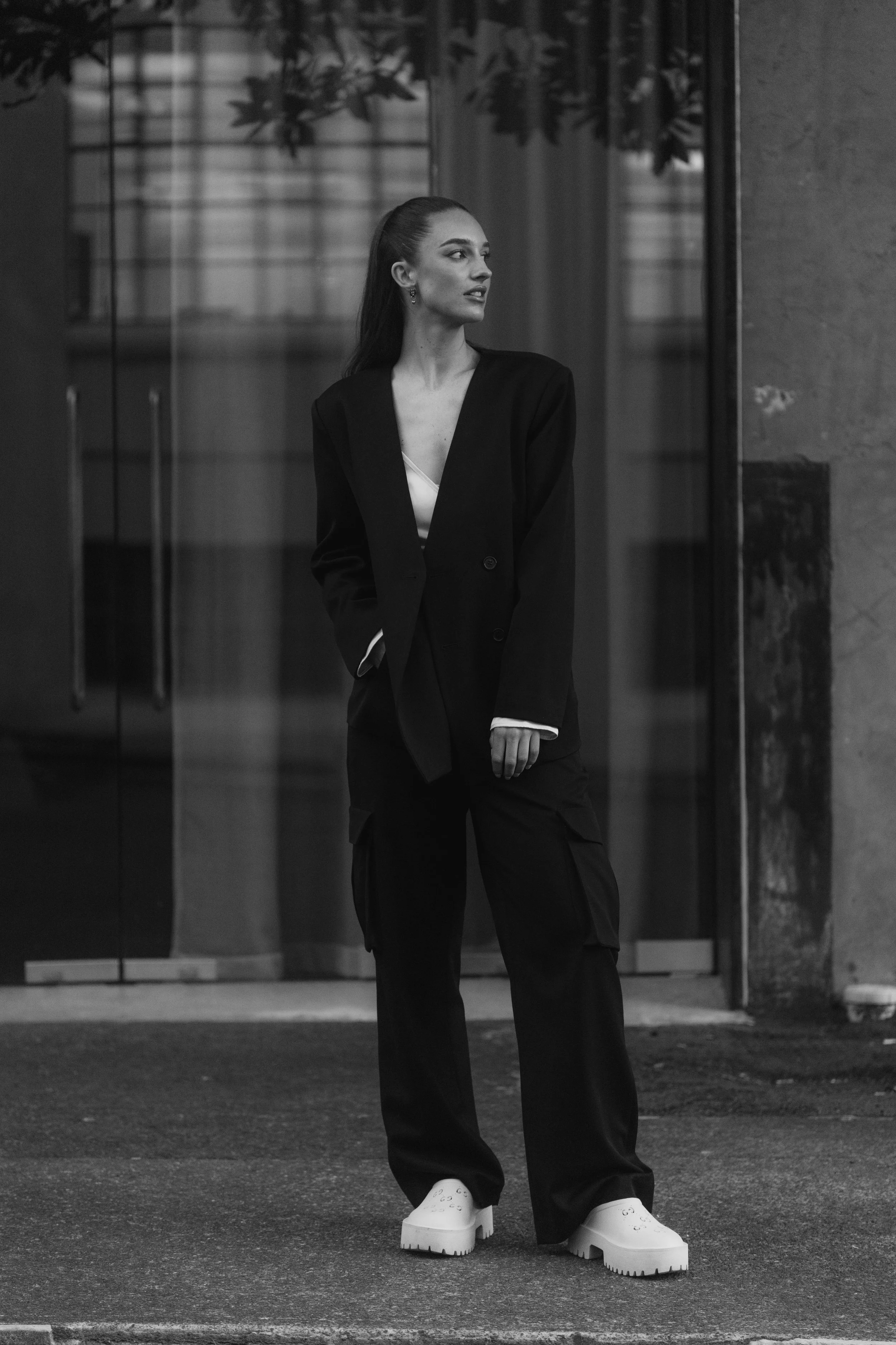 A woman standing outside near a building, wearing oversized shoes and a suit, looking to her right. The photo is in black and white.