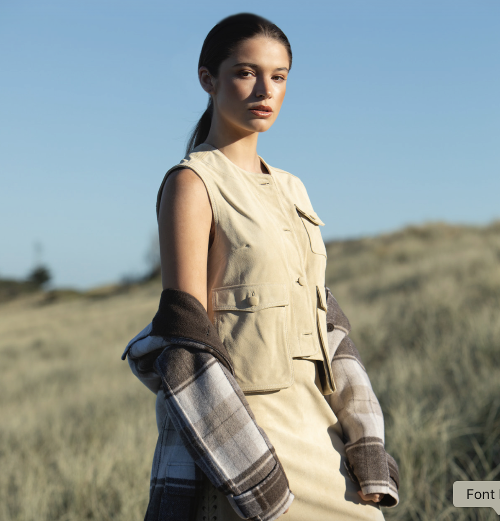A woman standing outdoors in a field of grass under a clear blue sky, wearing a sleeveless beige vest and a plaid shirt draped over her arms.