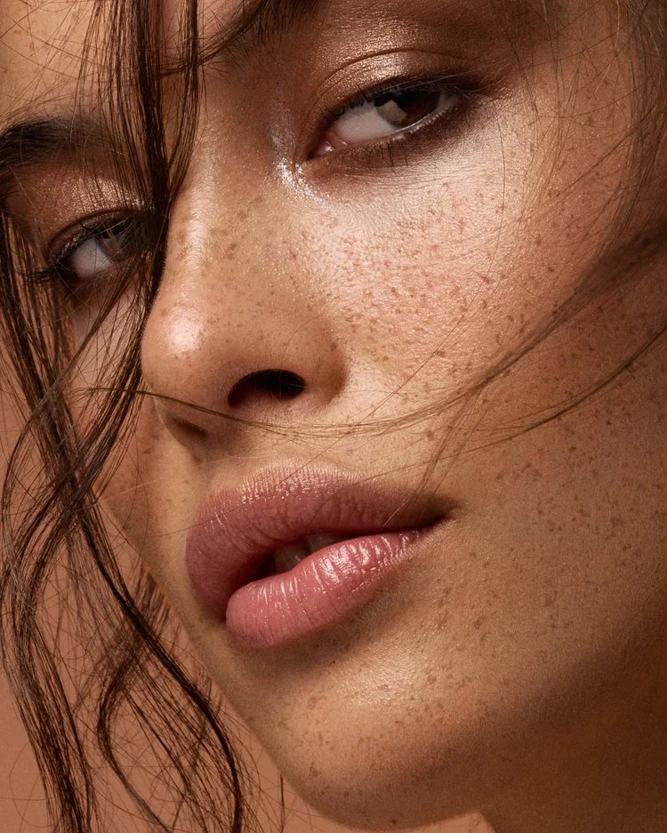 Close-up of a woman's face showing her skin with freckles, brown eyes, and lips with natural makeup, with loose hair strands.