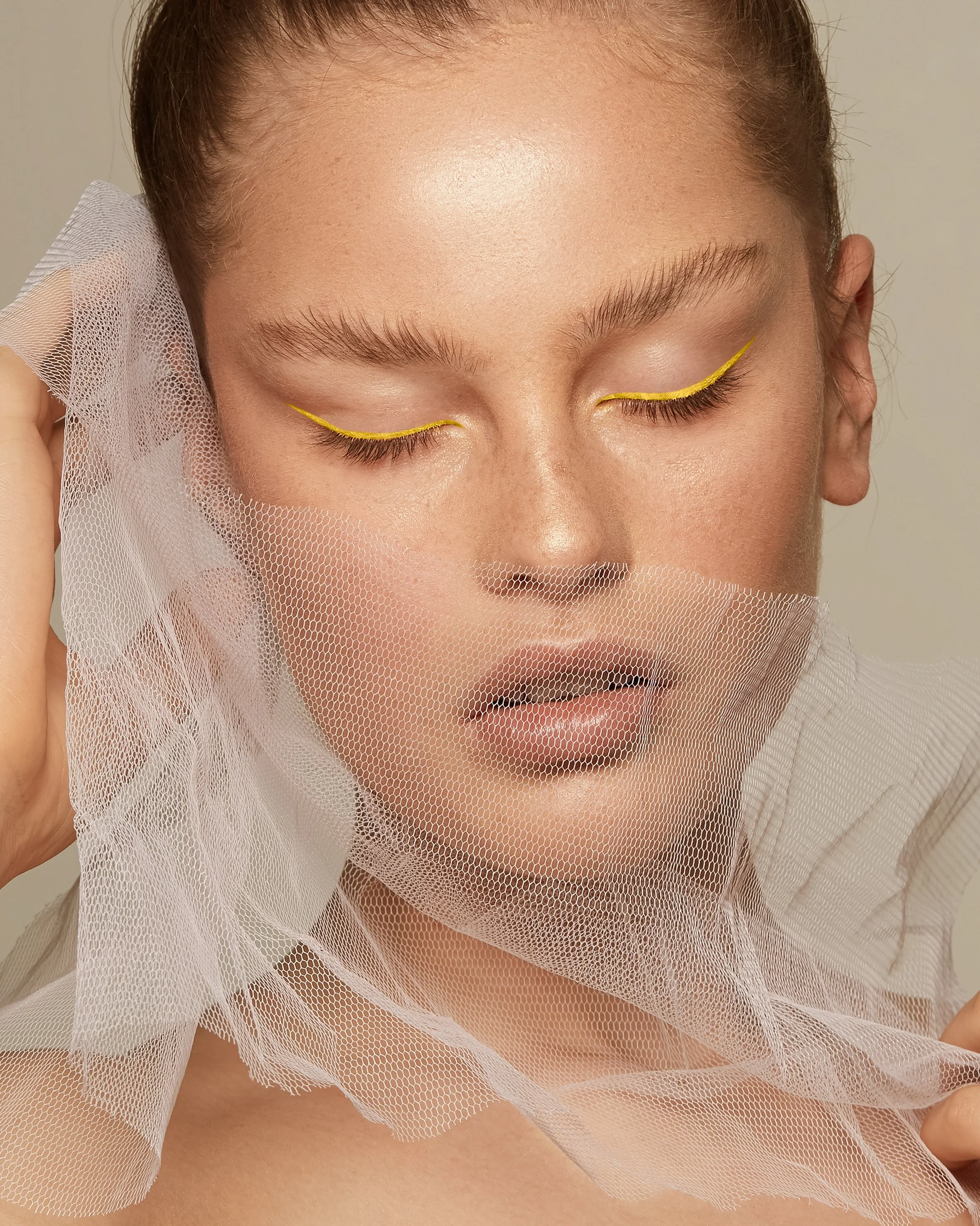 Close-up of a woman's face with closed eyes, wearing yellow eyeliner, and a sheer white veil on her head.
