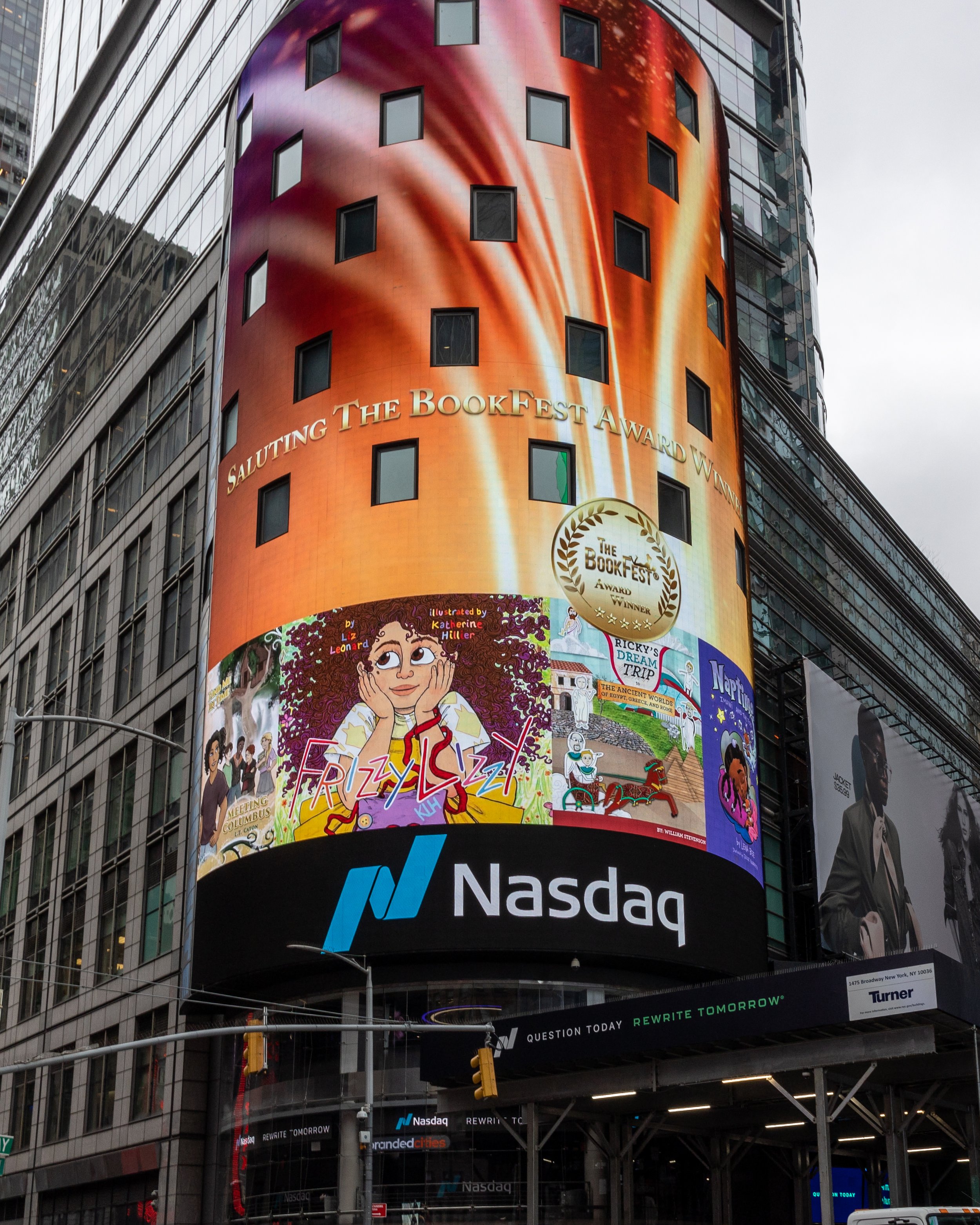 A tall building in New York City with a large digital billboard displaying colorful graphics, advertisements for a book festival, and the Nasdaq stock exchange logo at the bottom.