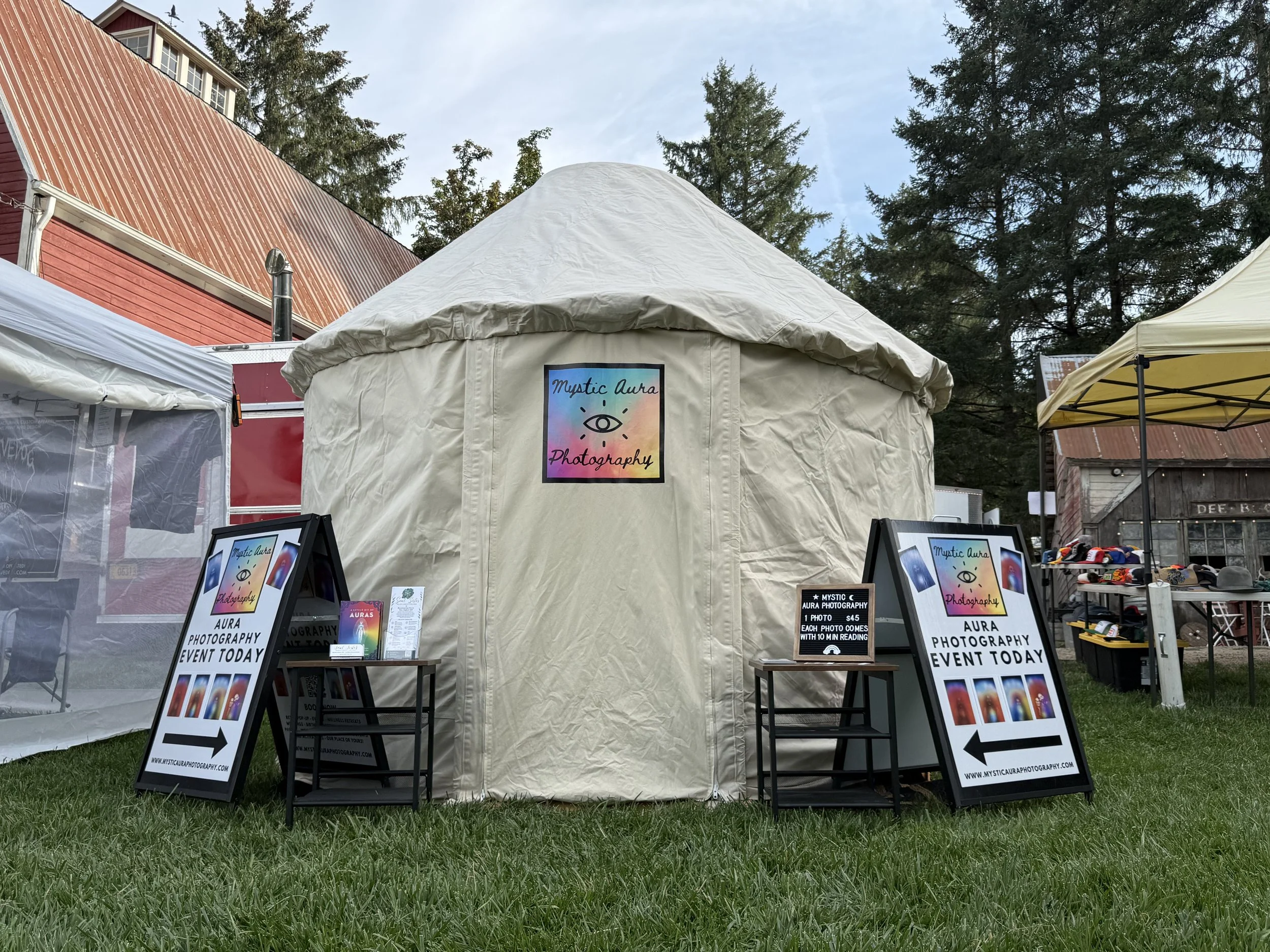 Outdoor event tent with signs advertising Mystic Aura Photography, set up on a grassy area with other tents and a red building in the background, surrounded by tall trees.