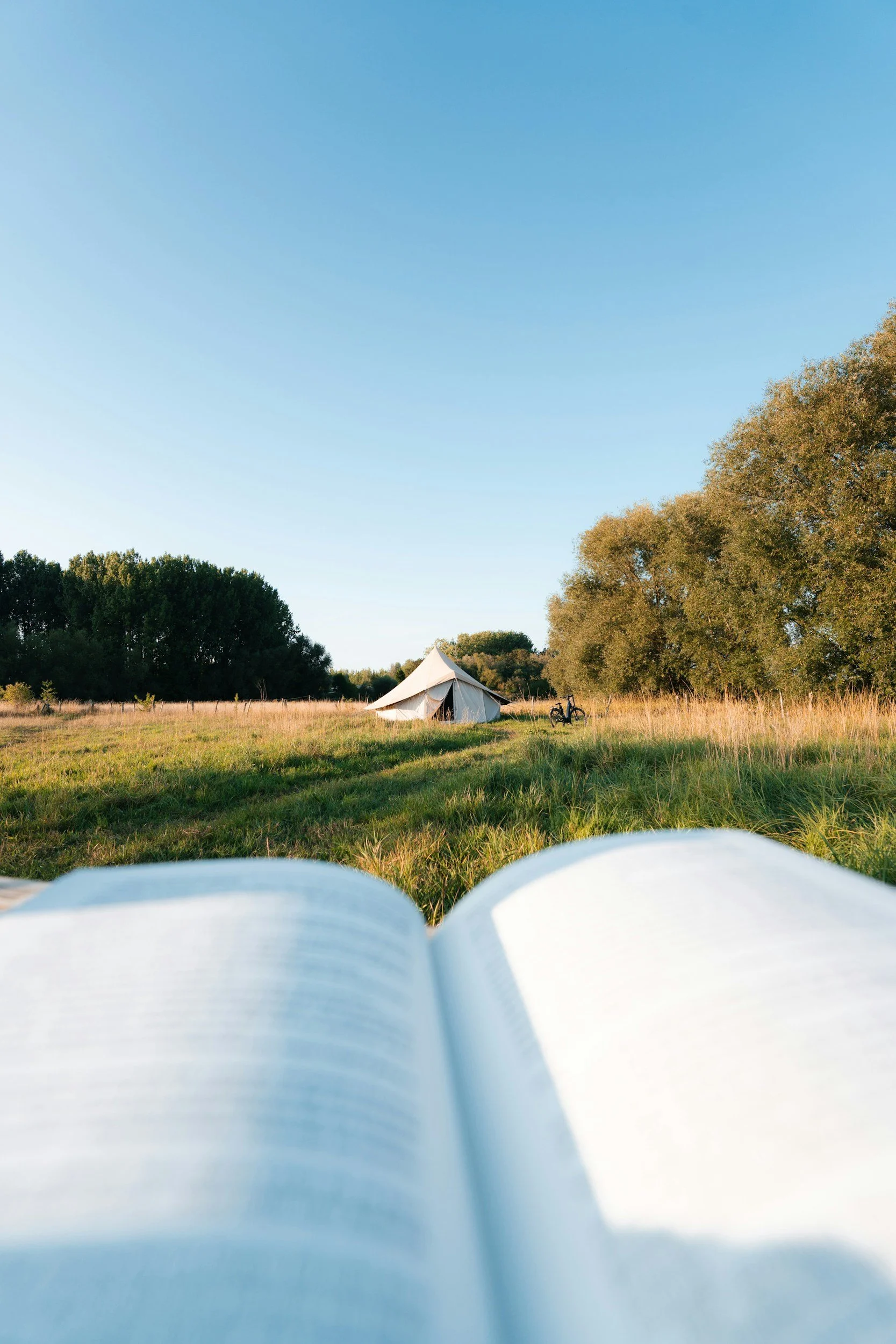 View of open book in the foreground, grassy field with trees, a tent, and a bicycle in the distance under a clear blue sky.