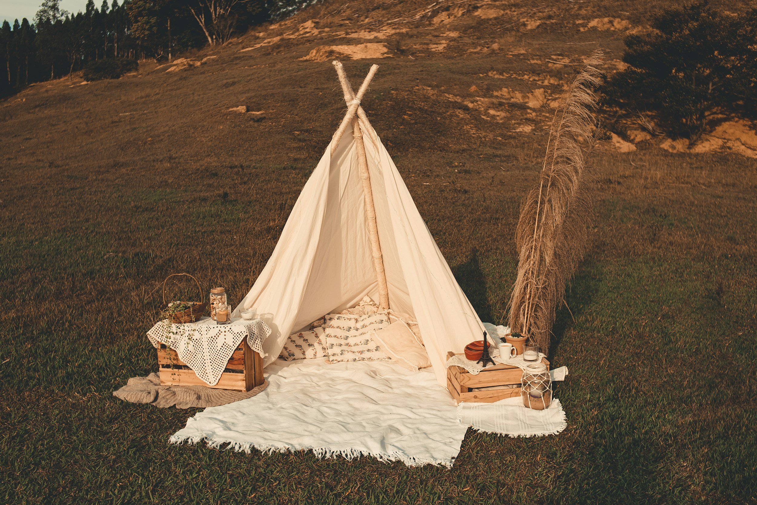 A cozy outdoor scene with a small cream-colored teepee tent set up on green grass in a natural landscape with hills and trees in the background, decorated with blankets, pillows, and various small items for a picnic or gathering.