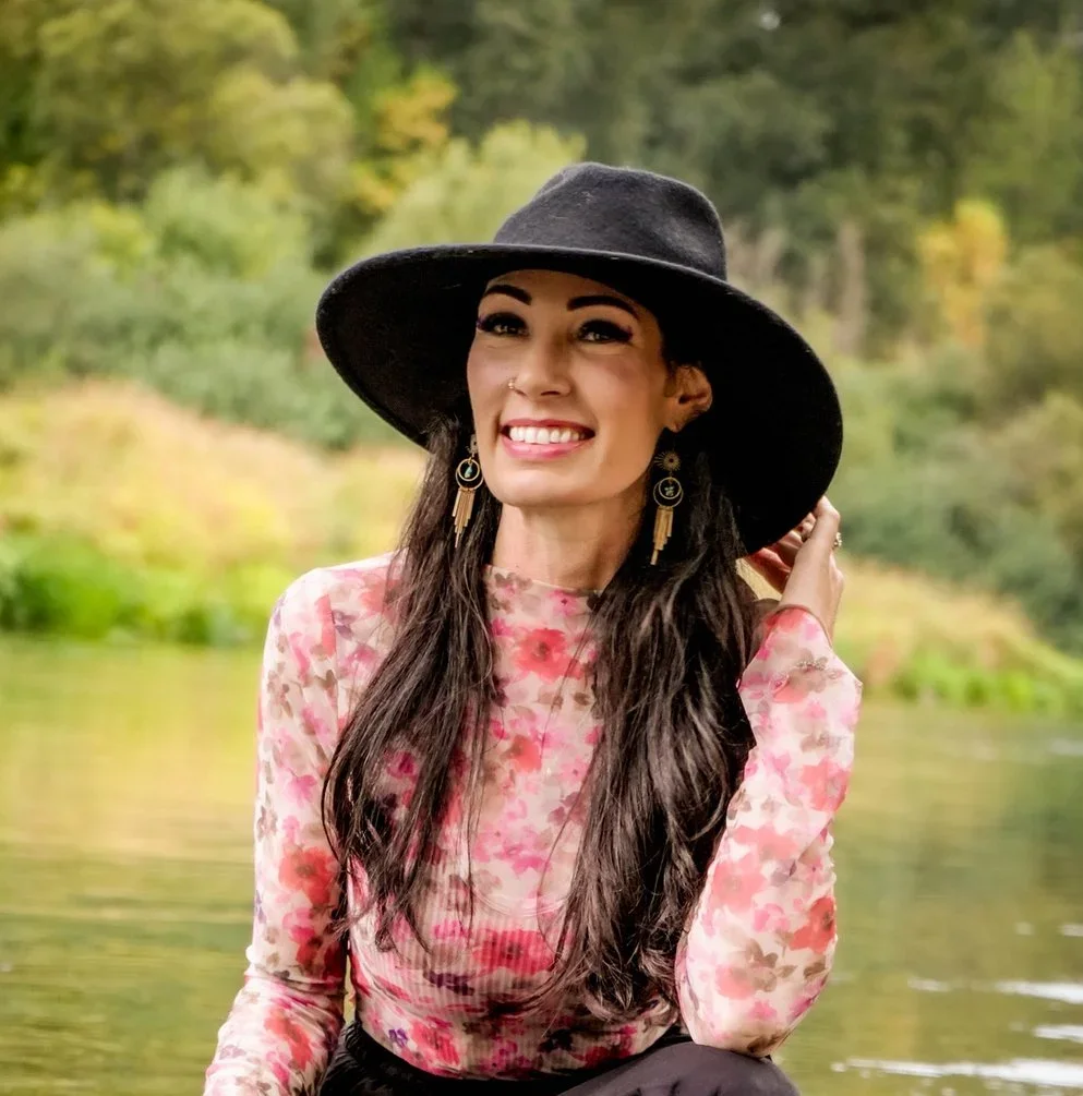 A woman with long dark hair, wearing a large black floppy hat and a pink floral blouse, smiling outdoors near a body of water with trees in the background.
