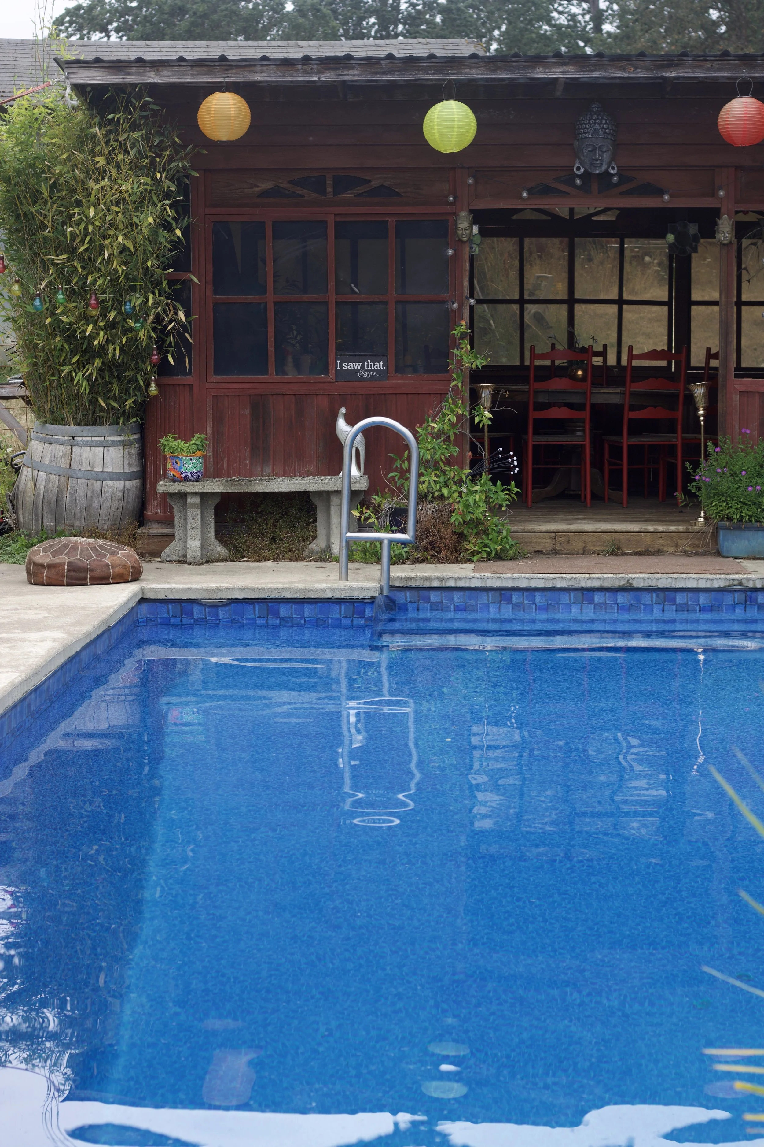 A backyard scene with a swimming pool in the foreground, a red wooden patio with potted plants and chairs, and hanging paper lanterns in yellow, green, and red. There are decorative masks on the patio wall and a stone bench with a small plant. A sign in the window reads 'I saw that.'