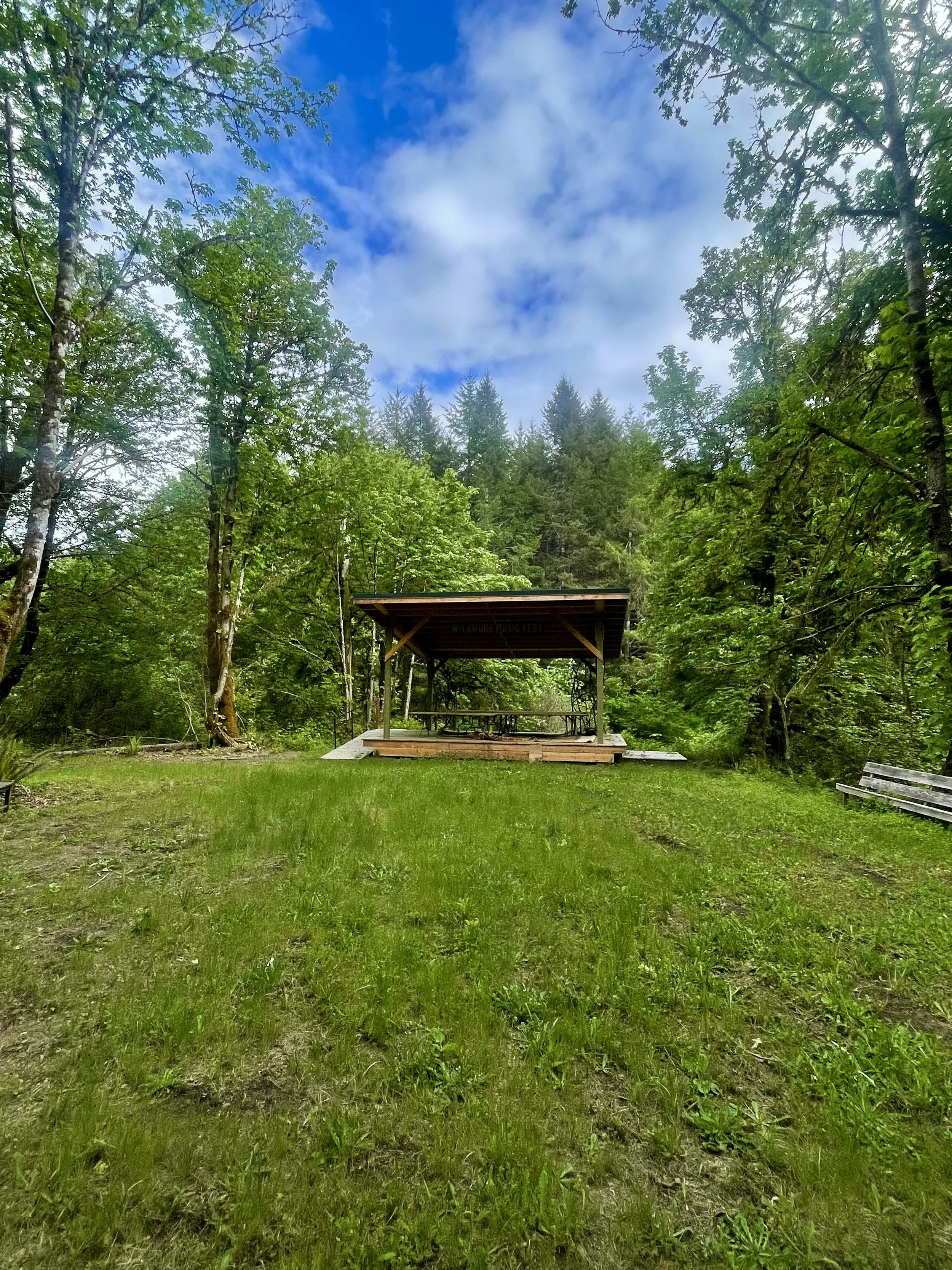 A small wooden stage with a roof and a picnic table underneath is set in a forest clearing surrounded by tall trees with green leaves. The sky is partly cloudy with patches of blue visible.
