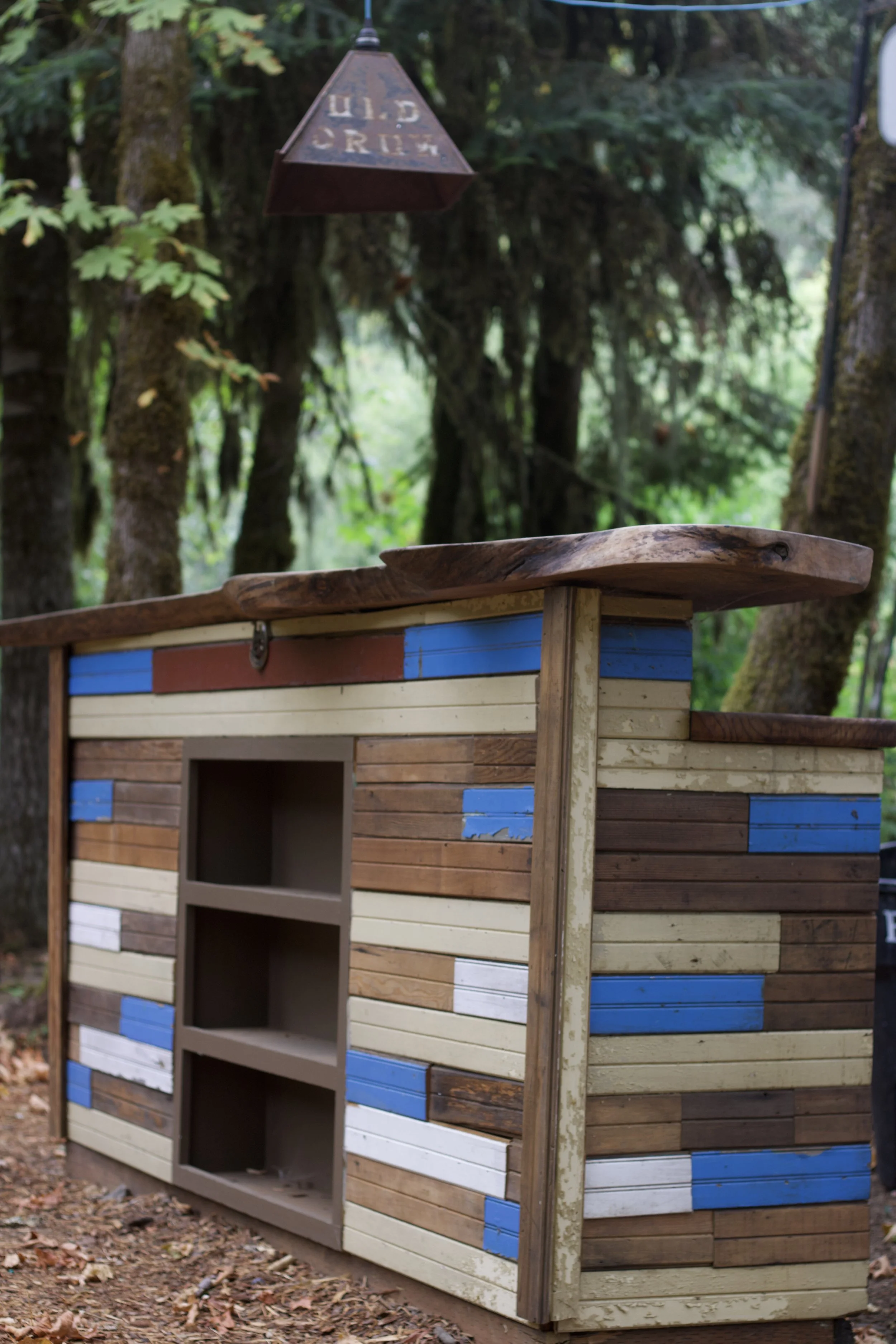 A colorful wooden small house with a window on the front, set in a wooded outdoor area.