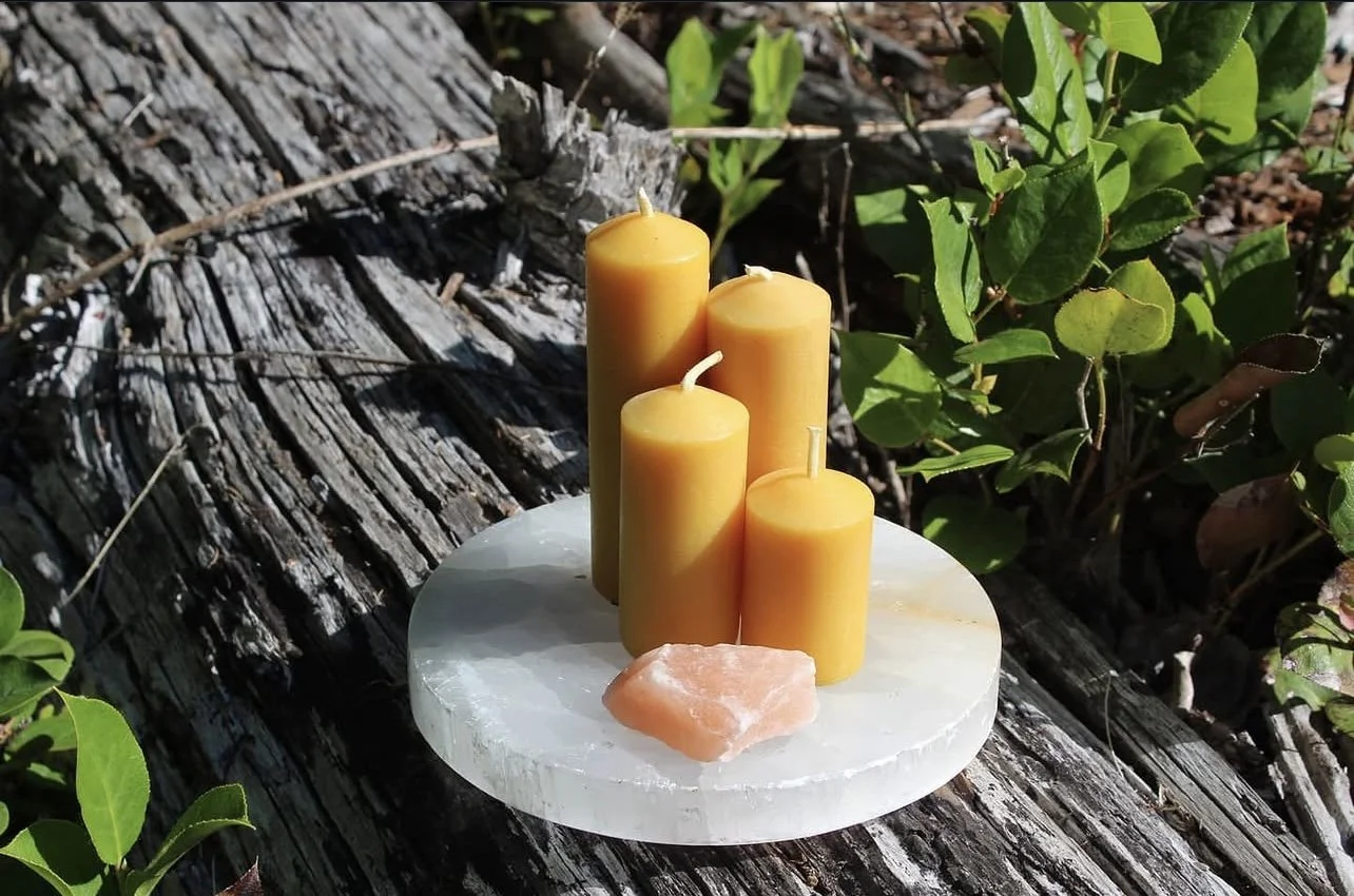 Five yellow candles of varying heights on a round white surface, with a pink salt crystal in front, outdoors on a weathered tree trunk with green foliage nearby.